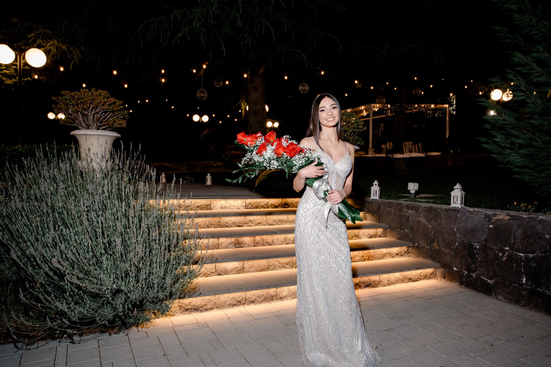 Ragazza in abito elegante argentato su una scalinata esterna illuminata di notte, con bouquet di rose rosse in primo piano. Sullo sfondo si vedono il giardino scuro, le luci calde appese tra gli alberi, grandi vasi decorativi e l'atmosfera scenografica dell'ingresso.