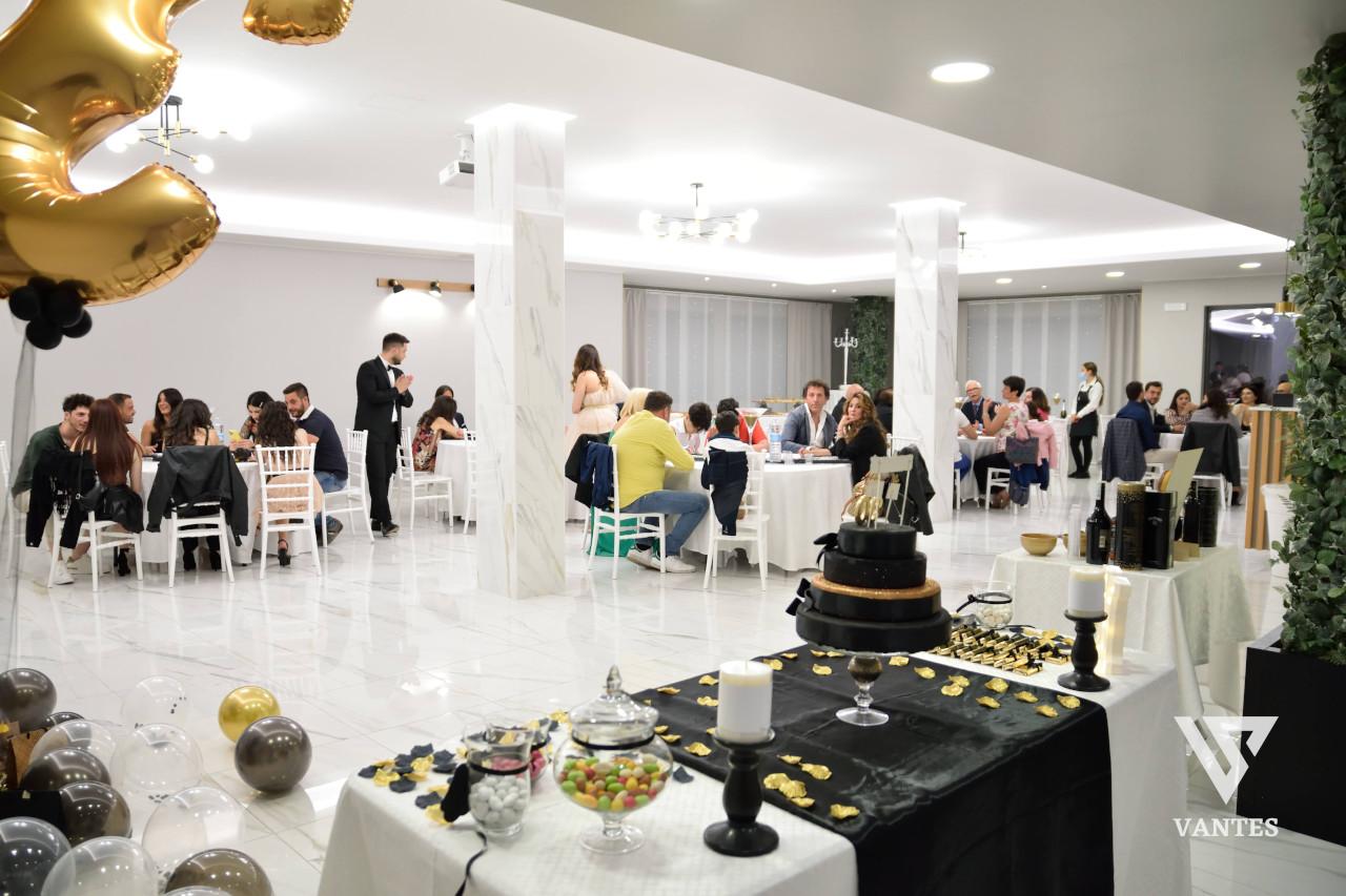 Sala evento luminosa con ospiti seduti ai tavoli rotondi durante una festa di compleanno, in un ambiente bianco e moderno con pilastri effetto marmo e pavimento lucido. In primo piano c'è un tavolo dessert nero e oro con torta a piani scura, dolci, bottiglie e palloncini metallici sparsi sul pavimento.