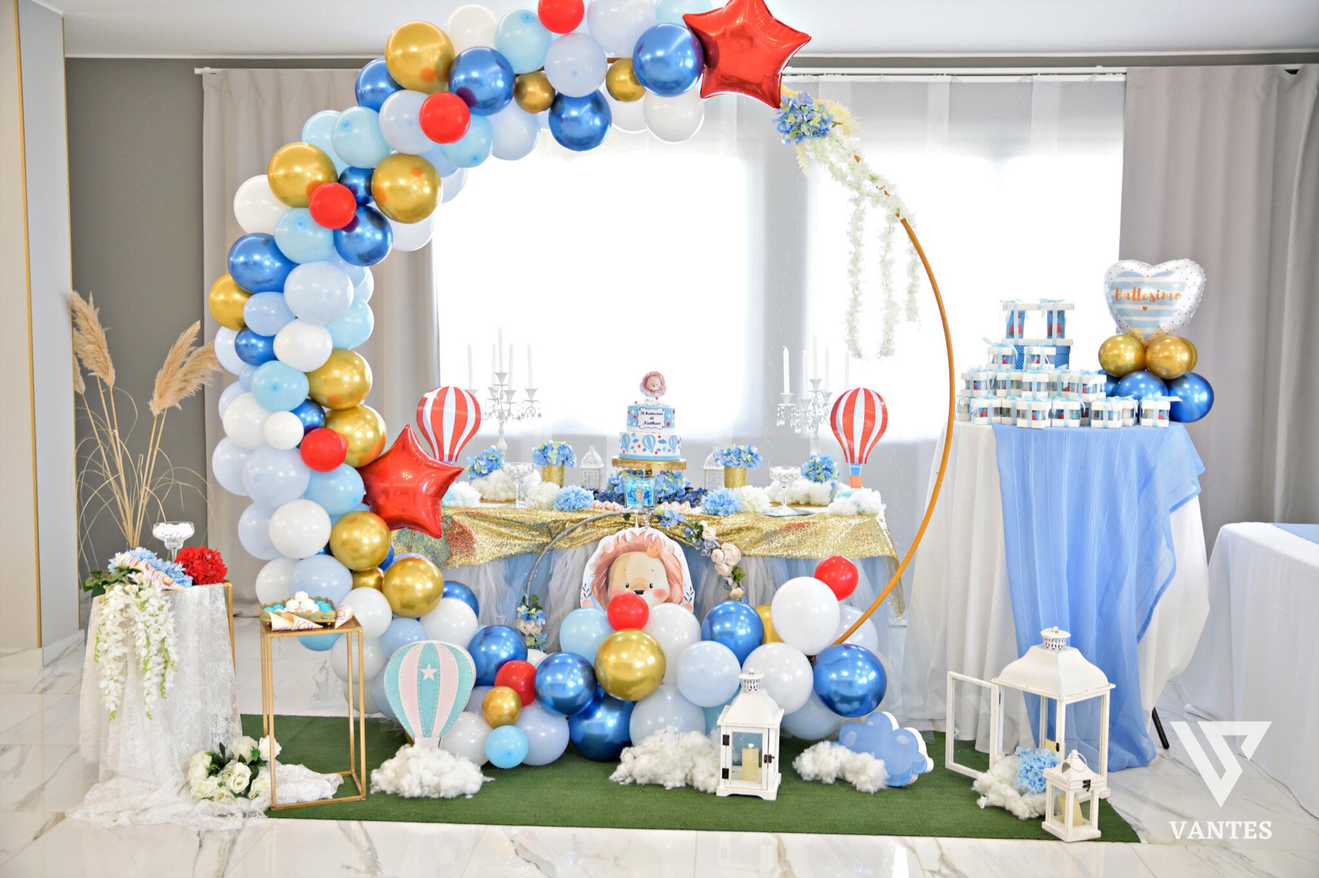 Allestimento scenografico per battesimo all'esterno o in sala, con grande arco di palloncini azzurri, bianchi, oro e rossi che incornicia il tavolo centrale. Sullo sfondo ci sono un tavolo dolci con confetti e bomboniere, elementi decorativi a tema baby, un palloncino a forma di cuore e grandi vasi floreali ai lati.
