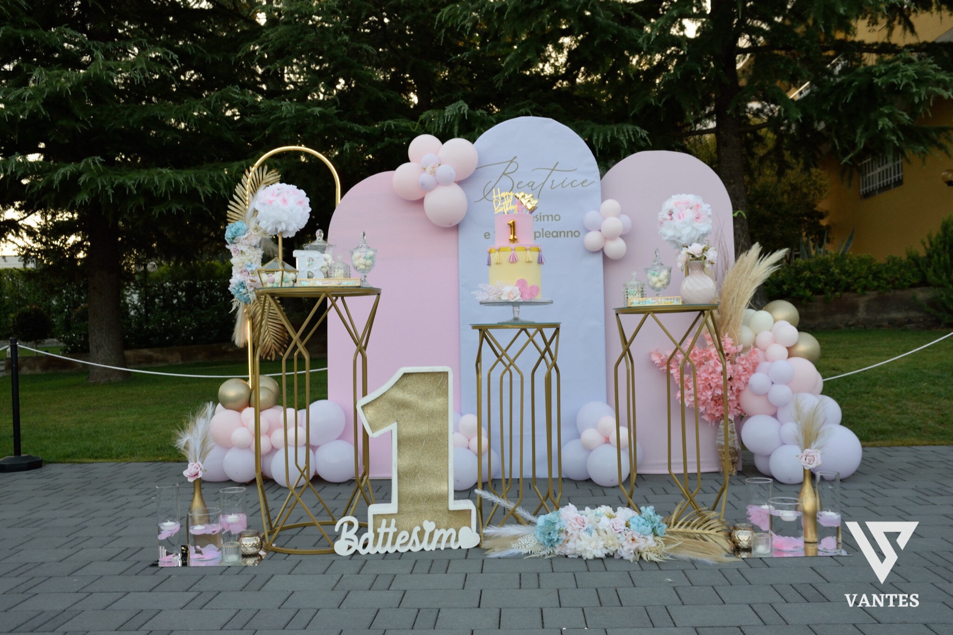 Allestimento esterno per battesimo o primo compleanno con fondale rosa e azzurro, nome della festeggiata in evidenza e un grande numero 1 dorato al centro. Attorno ci sono palloncini pastello, composizioni floreali, tavolini metallici con confettata e decorazioni scenografiche su prato e pavimentazione esterna.