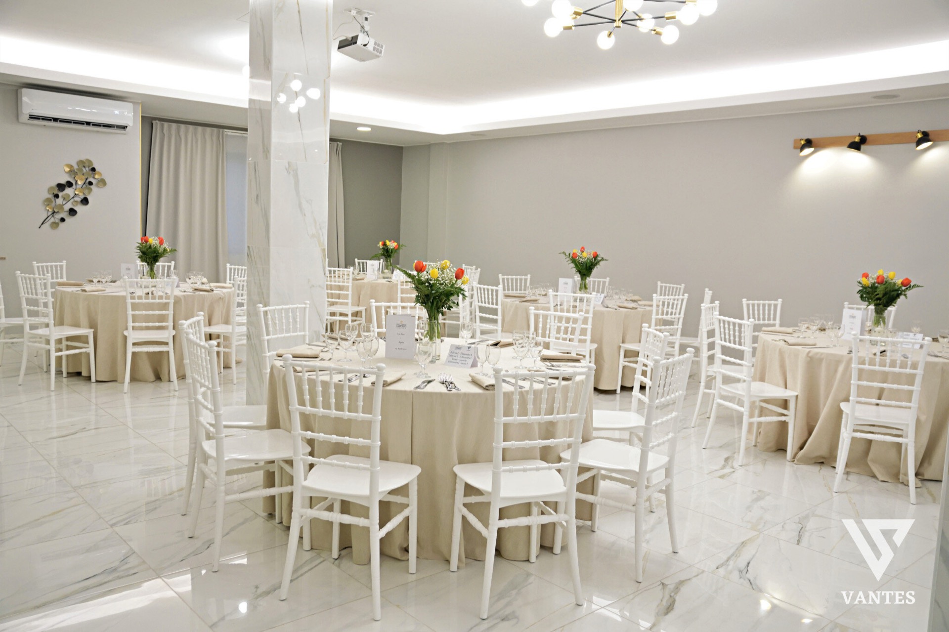 Sala interna con tavoli rotondi apparecchiati, tovaglie color sabbia, sedie bianche e piccoli centrotavola floreali su ogni tavolo. La scena mostra un allestimento ordinato e luminoso per il ricevimento, con pavimento chiaro effetto marmo e pareti neutre.