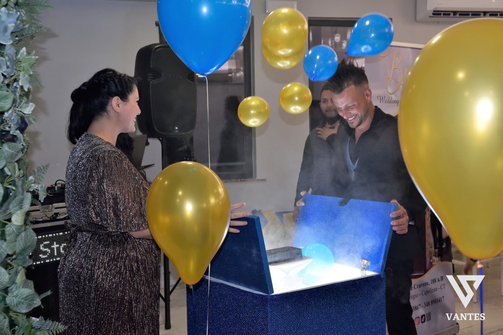 Scena di sorpresa con un grande box blu scintillante aperto al centro della sala, da cui esce fumo scenico e una luce azzurra interna. Intorno ci sono palloncini oro e blu, una donna di fronte al box e un uomo sorridente sulla destra, in un momento di festa molto teatrale.