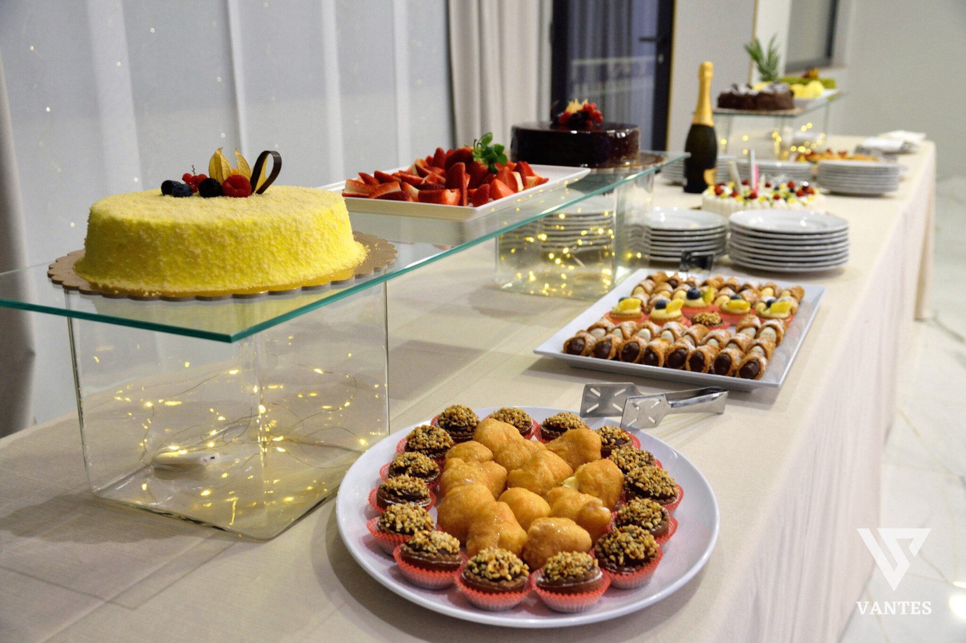 Buffet dei dolci allestito su un lungo tavolo con torte, frutta fresca, pasticcini e vassoi di dessert disposti su più livelli. In primo piano si vedono ciambelle glassate e piccoli dolci al cioccolato, mentre sul fondo ci sono piatti impilati, una bottiglia di spumante e altri vassoi da servizio.