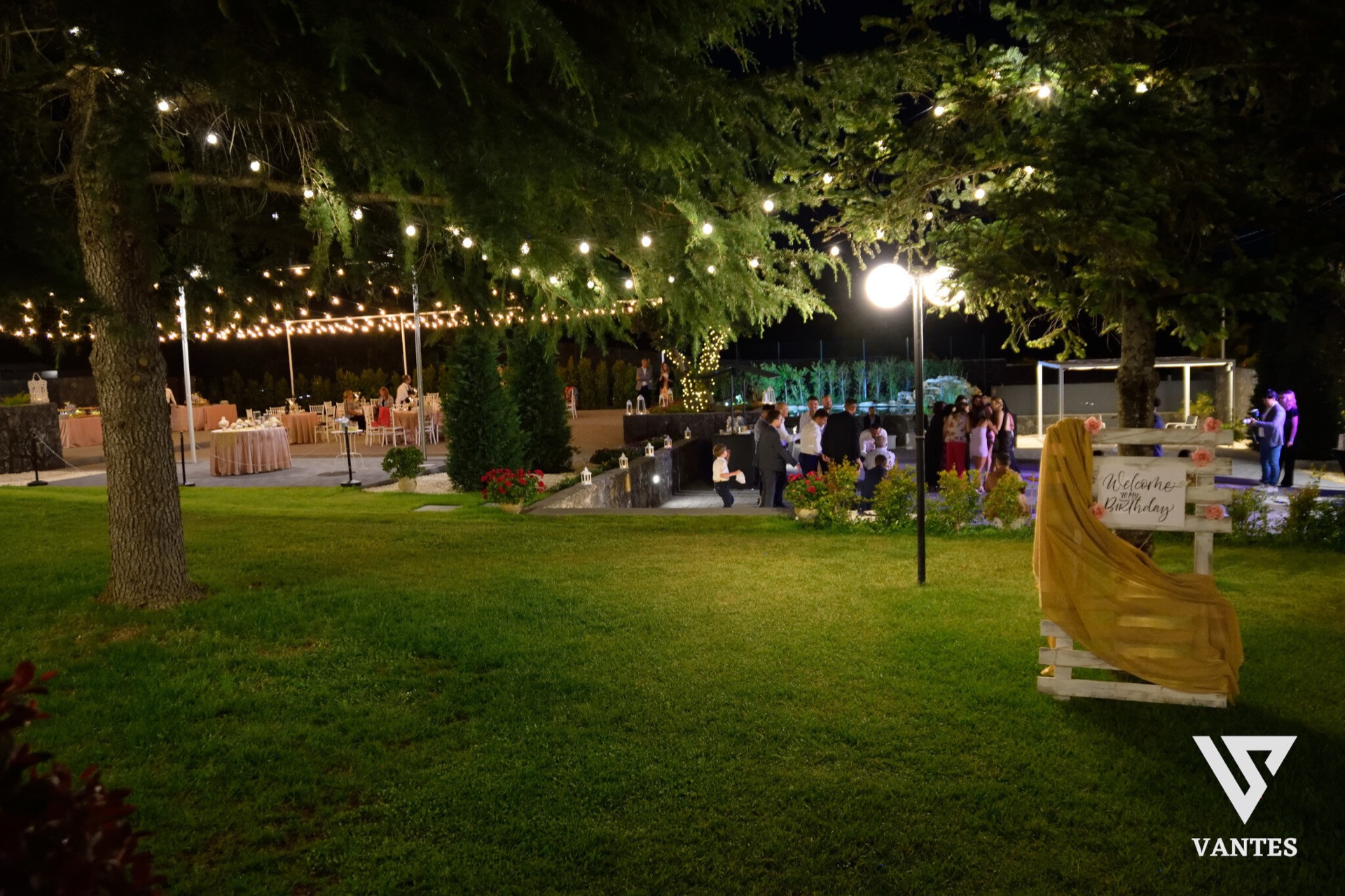 Festa di compleanno serale in giardino con prato in primo piano, tavoli apparecchiati sullo sfondo, gruppi di ospiti vicino al percorso centrale e luci calde sospese tra gli alberi. In evidenza c'è anche un cartello di benvenuto 'Welcome to my Birthday' accanto all'ingresso decorato.