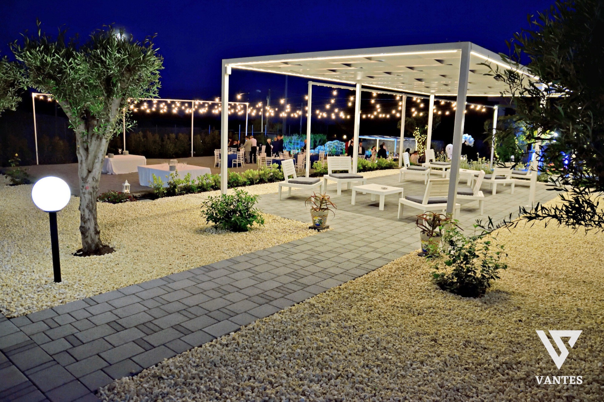Lounge esterna serale nel giardino della villa con percorso in pietra, ghiaia chiara, sedute bianche sotto la pergola illuminata e piccoli gruppi di ospiti sullo sfondo. Le luci calde sospese tra gli alberi e le aiuole curate danno alla scena un'atmosfera elegante e rilassata.