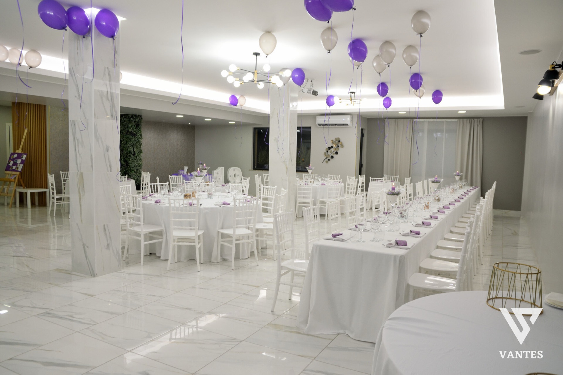 Sala interna luminosa con tavoli rotondi e una lunga tavolata apparecchiata, sedie bianche e piccoli centrotavola viola. Dal soffitto scendono palloncini viola e bianchi, mentre i pilastri effetto marmo e il pavimento lucido danno un aspetto elegante e ordinato all'allestimento.
