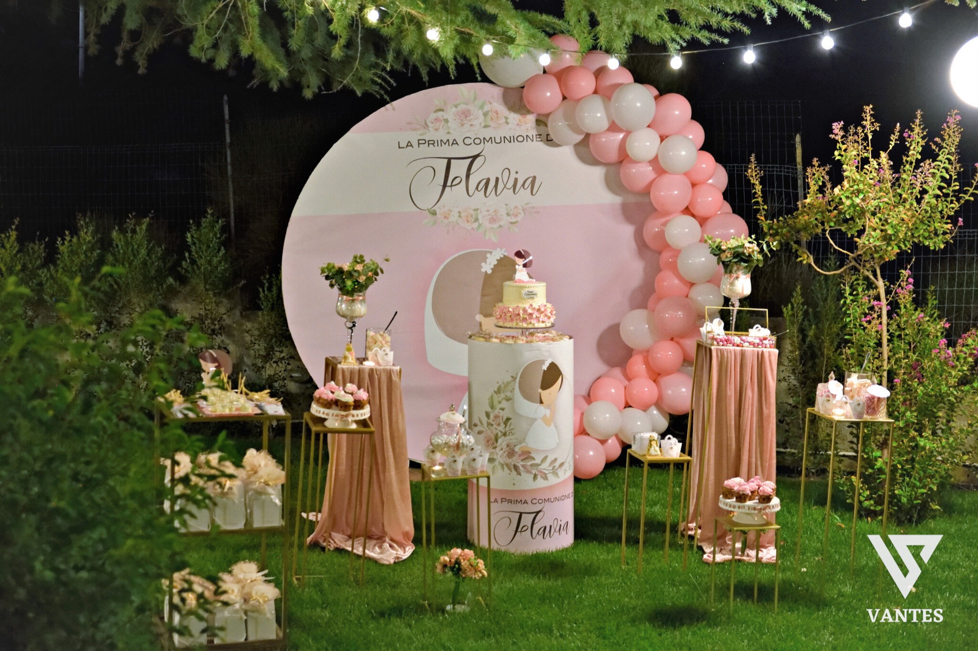 Allestimento esterno per la Prima Comunione con grande fondale rosa, arco di palloncini rosa e bianchi, nome della festeggiata in evidenza e torta scenografica al centro. Intorno ci sono tavolini dorati con confettata, dolci e bomboniere, disposti su prato verde sotto luci calde serali.