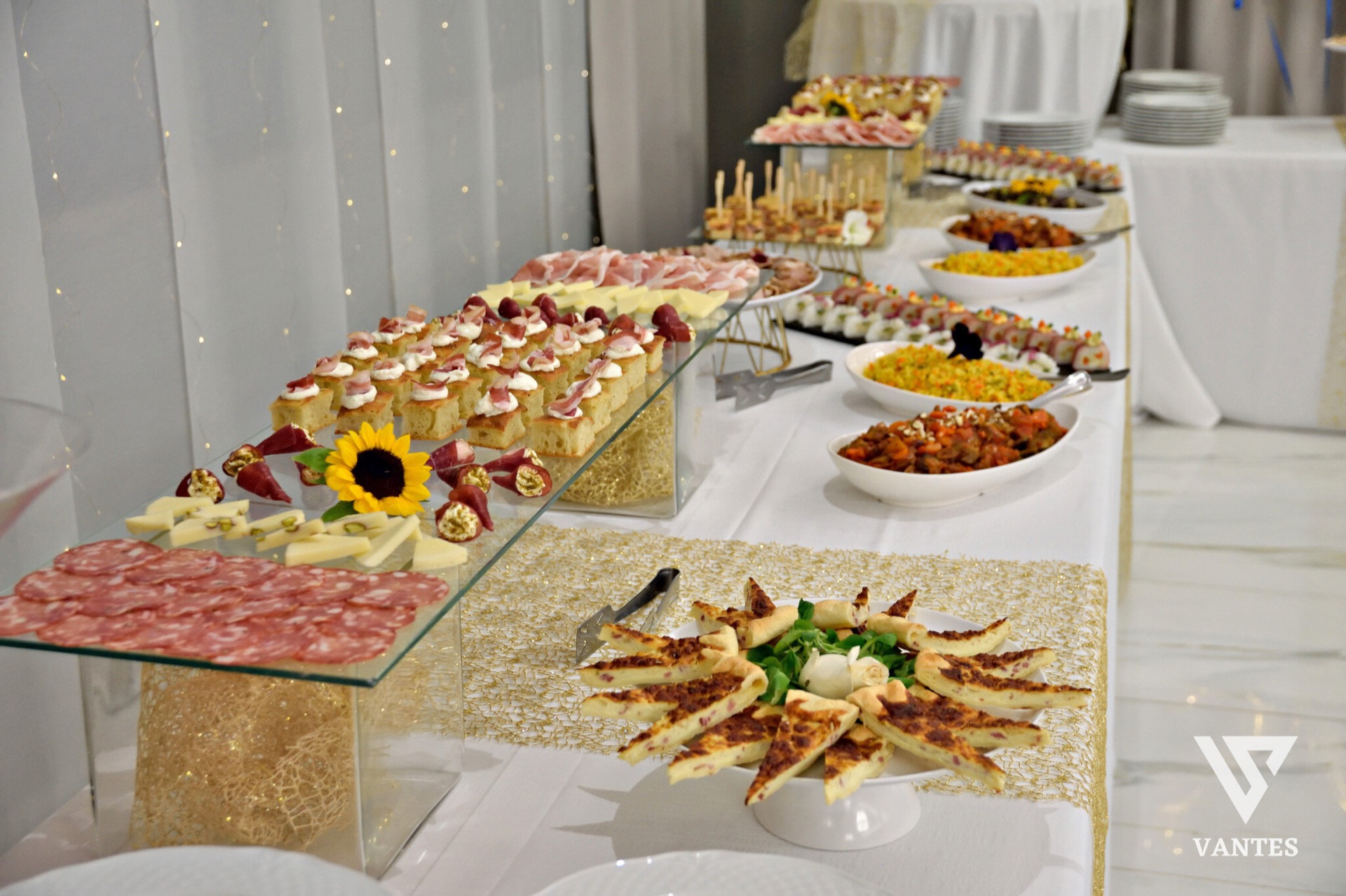 Lungo buffet salato allestito su più livelli con salumi, formaggi, tartine, spiedini, piccole portate calde e vassoi di antipasti disposti in file ordinate. Sul fronte ci sono piatti decorativi con assaggi a spicchi, mentre sullo sfondo si vedono piatti impilati e una tovaglia bianca con dettagli dorati.