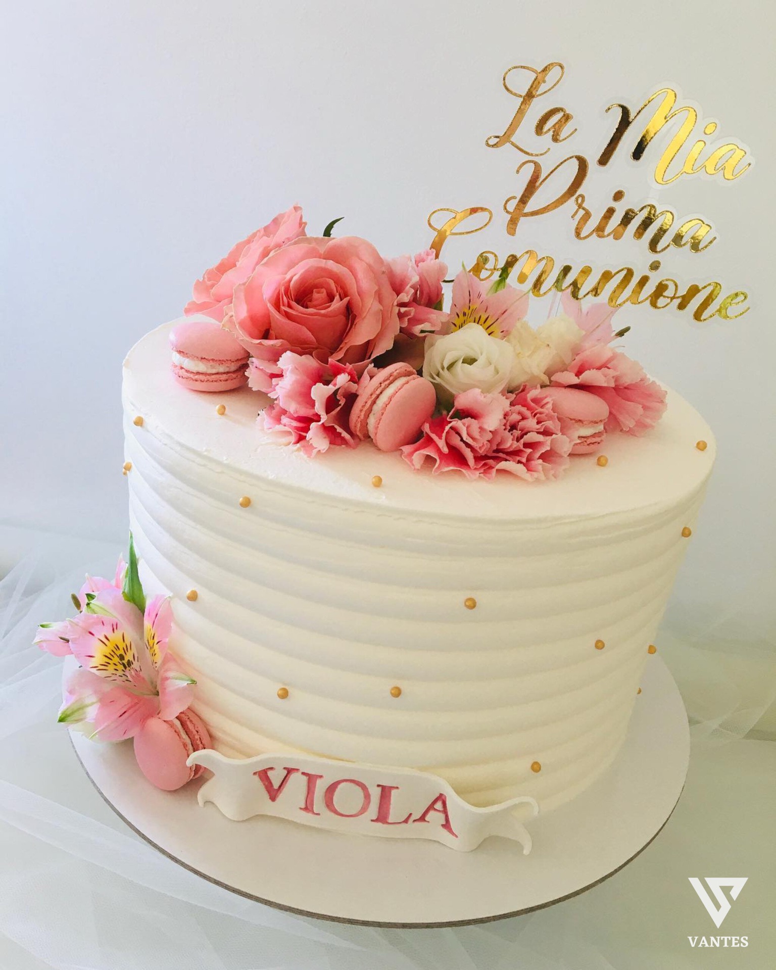 Torta bianca a piani alti decorata con fiori rosa, macarons rosa, piccoli dettagli dorati e scritta 'La Mia Prima Comunione' in oro sul topper. Sul fronte della base compare il nome 'VIOLA' in rosa, in una composizione elegante e delicata da tavolo torta.