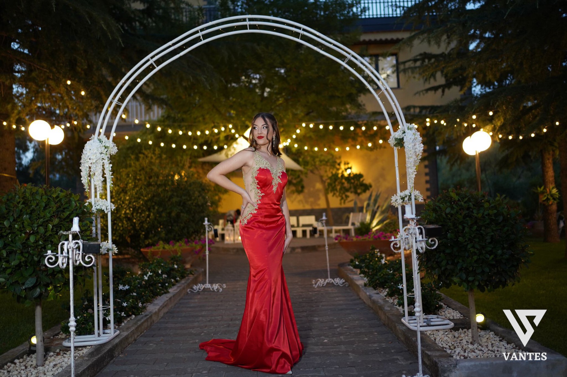 Ragazza in abito lungo rosso con dettagli dorati che posa al centro di un viale esterno decorato con un grande arco bianco, candele e fiori ai lati. Sullo sfondo si vedono le luci calde del giardino serale, i cespugli curati e la facciata della villa in penombra.