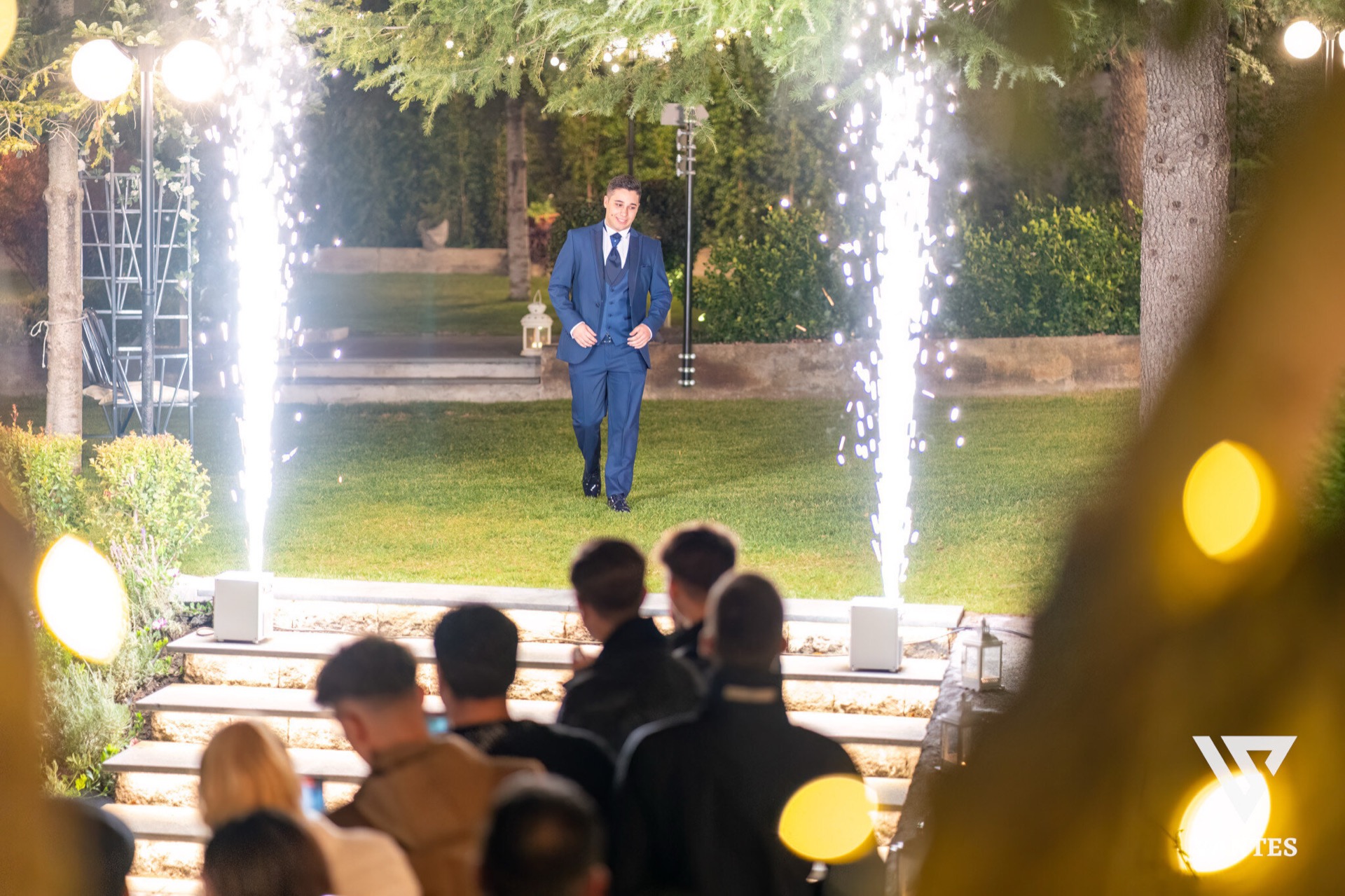 Ingresso scenografico notturno sul prato con due fontane luminose ai lati e un ragazzo in abito blu che avanza al centro dei gradini illuminati. In primo piano si vedono gli ospiti seduti e le luci calde del giardino, mentre il resto della scena è incorniciato da alberi e siepi curate.