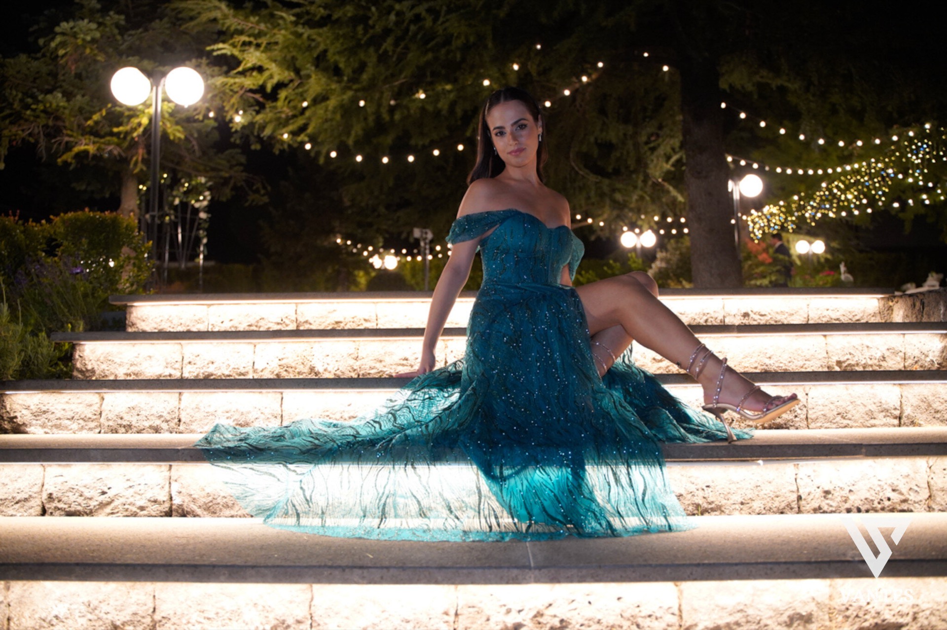 Festeggiata in abito lungo turchese seduta sulla scalinata esterna illuminata di notte, con il vestito che si apre ampio sui gradini. Sullo sfondo si vedono le luci calde del giardino, gli alberi e i lampioni che rendono la scena molto scenografica.
