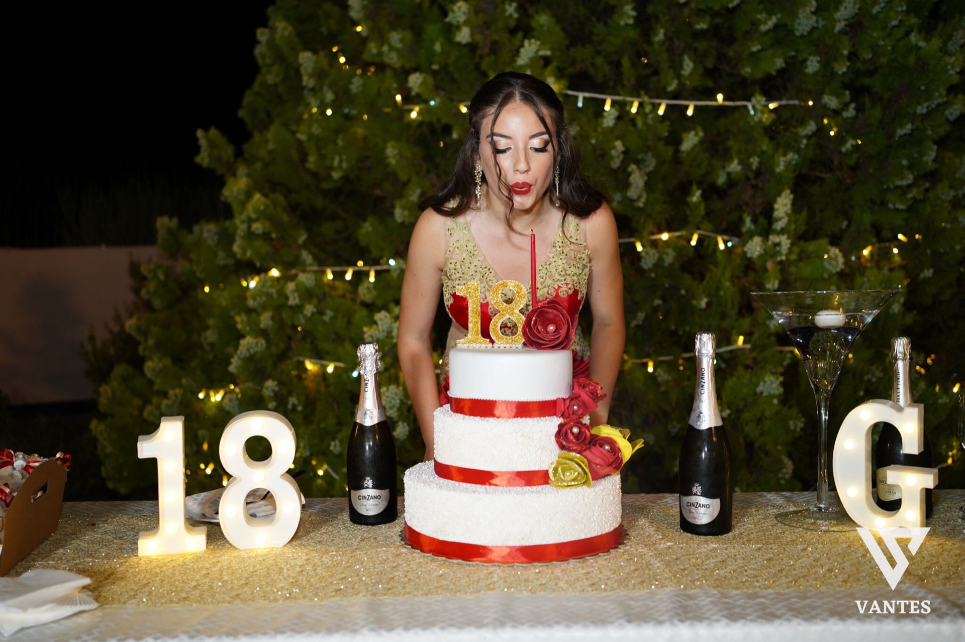 Tavolo torta all'aperto di notte con festeggiata in abito rosso e oro che soffia sulle candeline davanti a una torta a tre piani bianca e rossa. Sul tavolo ci sono numeri luminosi 18, bottiglie di spumante, un bicchiere cocktail a coppa e decorazioni floreali rosse e gialle su sfondo di siepe illuminata.