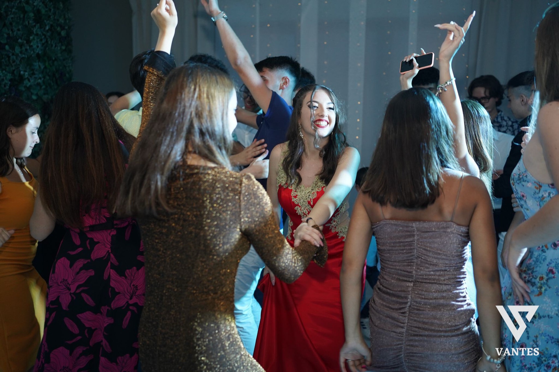Gruppo di ragazzi e ragazze che balla in sala attorno alla festeggiata in abito rosso, con le mani alzate e i volti illuminati da luci fredde e calde miste. Sullo sfondo si intravedono tende chiare, piccoli punti luce decorativi e l'atmosfera energica della festa danzante.