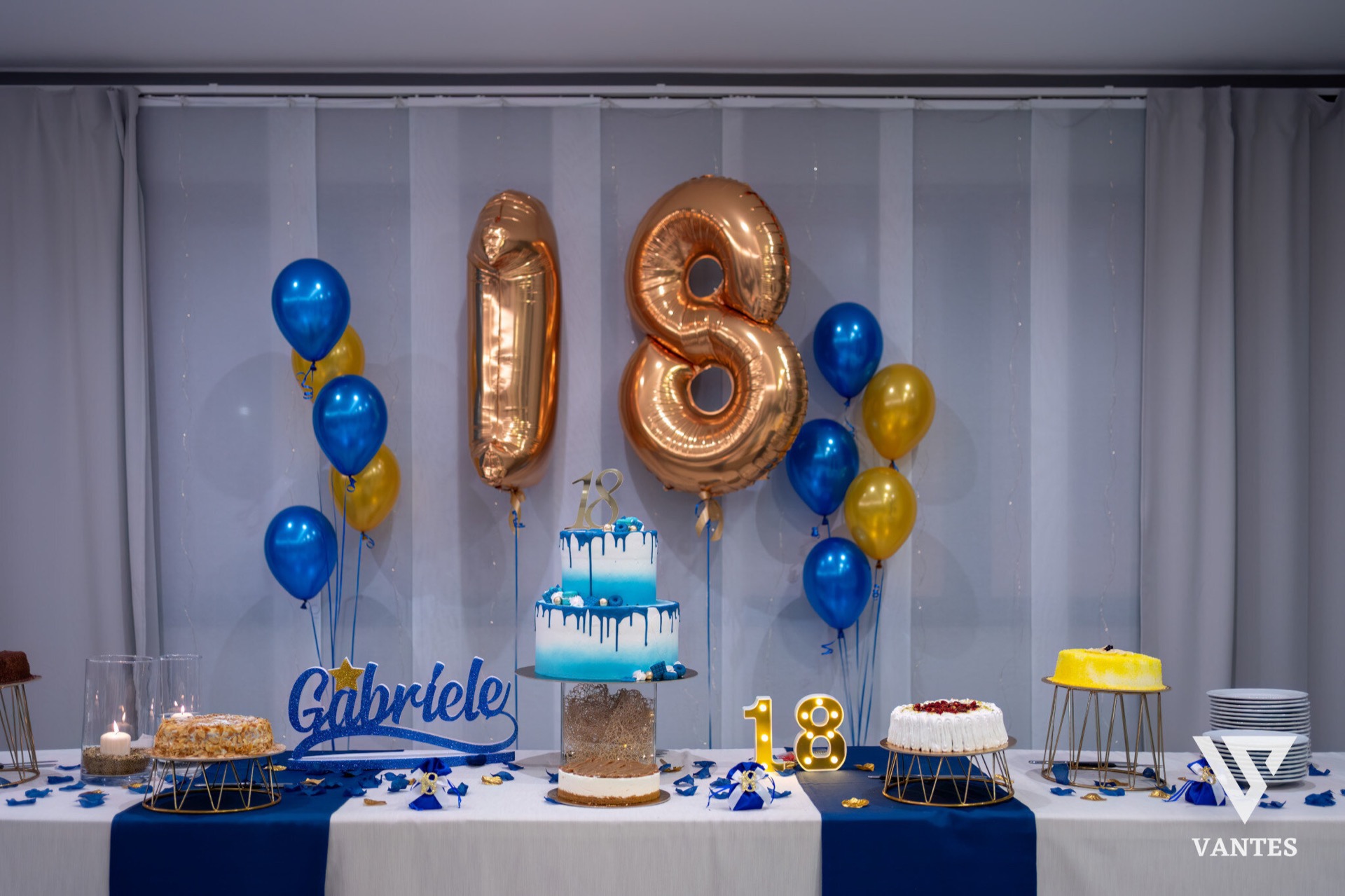 Tavolo torta per il diciottesimo con grande fondale di palloncini oro '18', nome 'Gabriele' in blu e due torte decorate in azzurro e bianco. Sul tavolo ci sono dolci monoporzione, candele, piatti impilati e coriandoli blu e oro distribuiti su tutta la superficie.