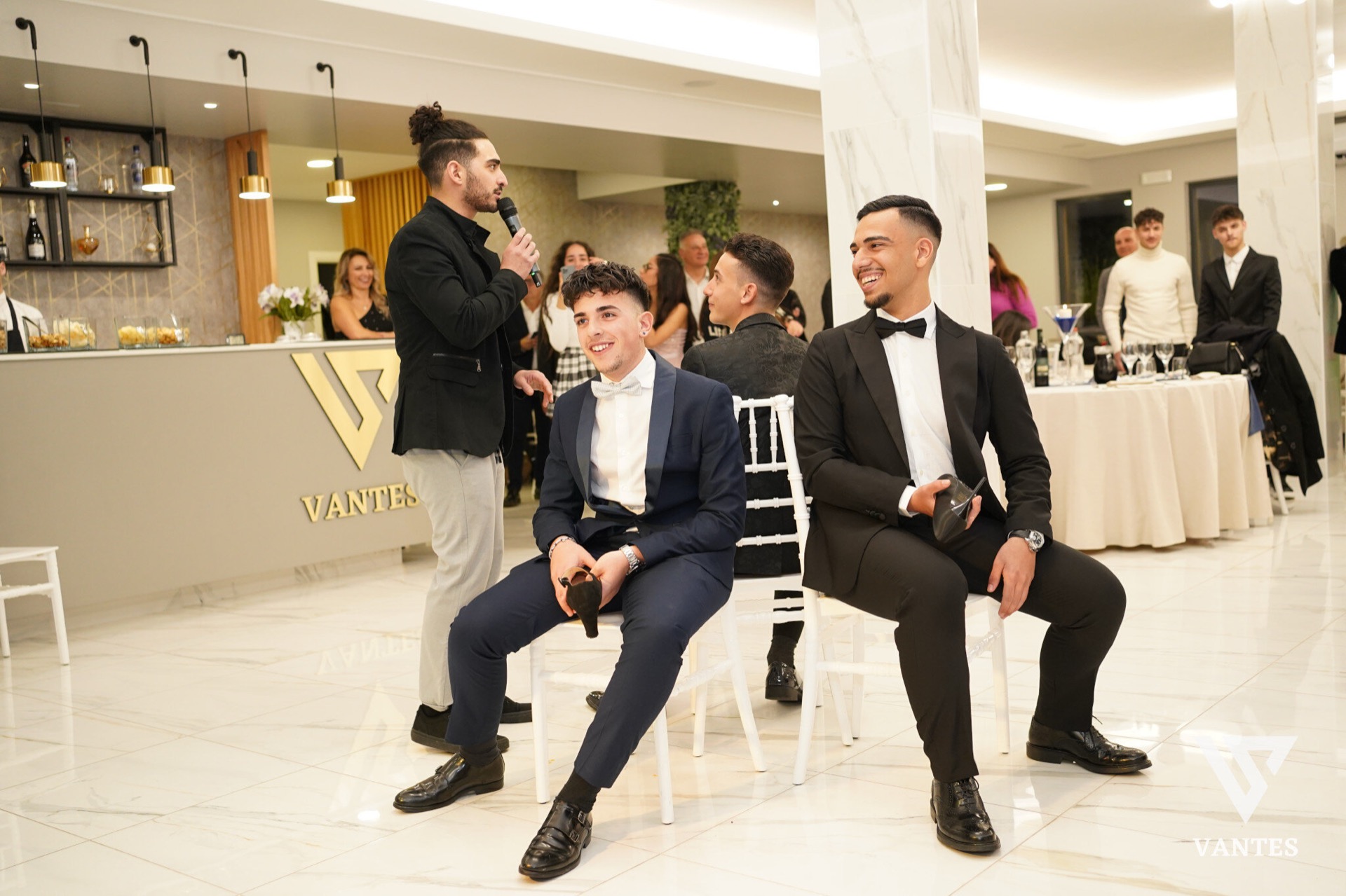 Momento in sala con un ragazzo che parla al microfono vicino al bancone e due giovani seduti in primo piano con abiti eleganti da cerimonia. Sullo sfondo ci sono altri ospiti, tavoli rotondi con tovaglie chiare e il banco bar illuminato, in un'atmosfera formale da ricevimento.