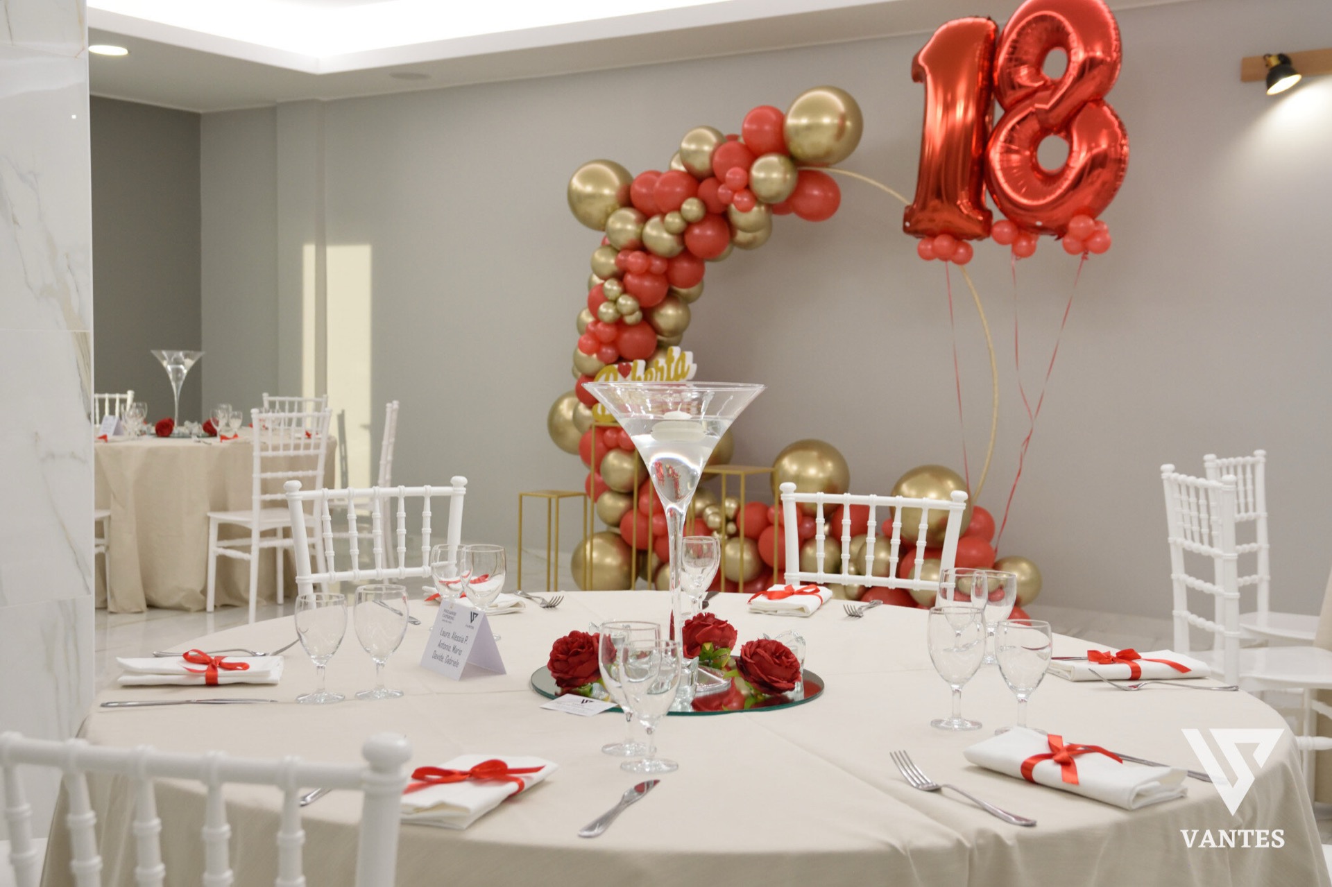 Tavolo rotondo apparecchiato con tovaglia chiara, calici, tovaglioli con nastri rossi e centrotavola floreale rosso al centro. Sullo sfondo si vede un arco di palloncini rosso e oro con numero 18 metallico, insieme ad altri tavoli coordinati nella sala.