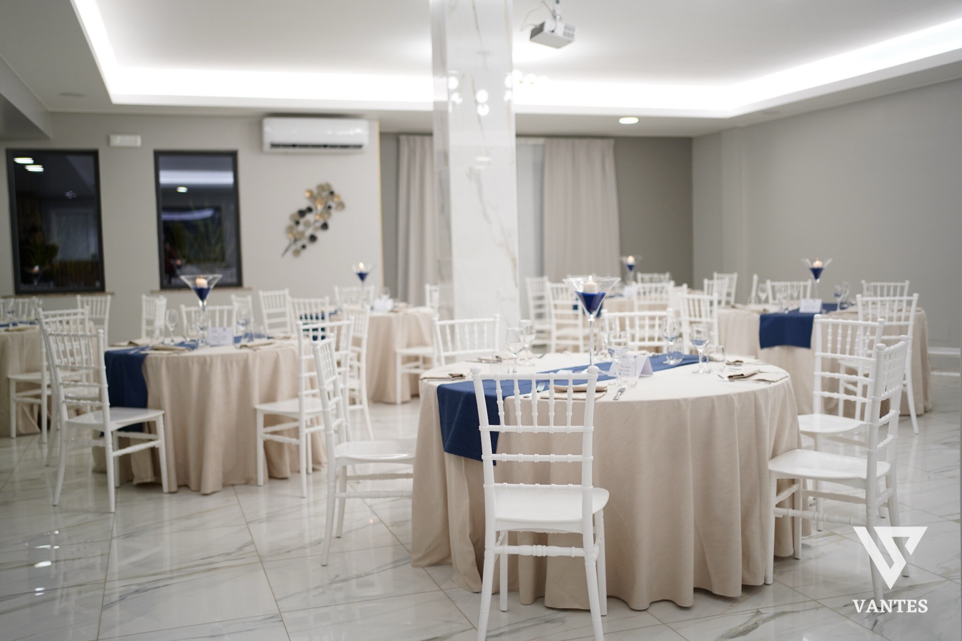 Sala interna luminosa con tavoli rotondi apparecchiati, tovaglie beige, runner blu e sedie bianche disposte in modo ordinato. Il pavimento lucido effetto marmo, i pilastri chiari e i dettagli minimalisti creano un ambiente elegante e sobrio per il ricevimento di laurea.