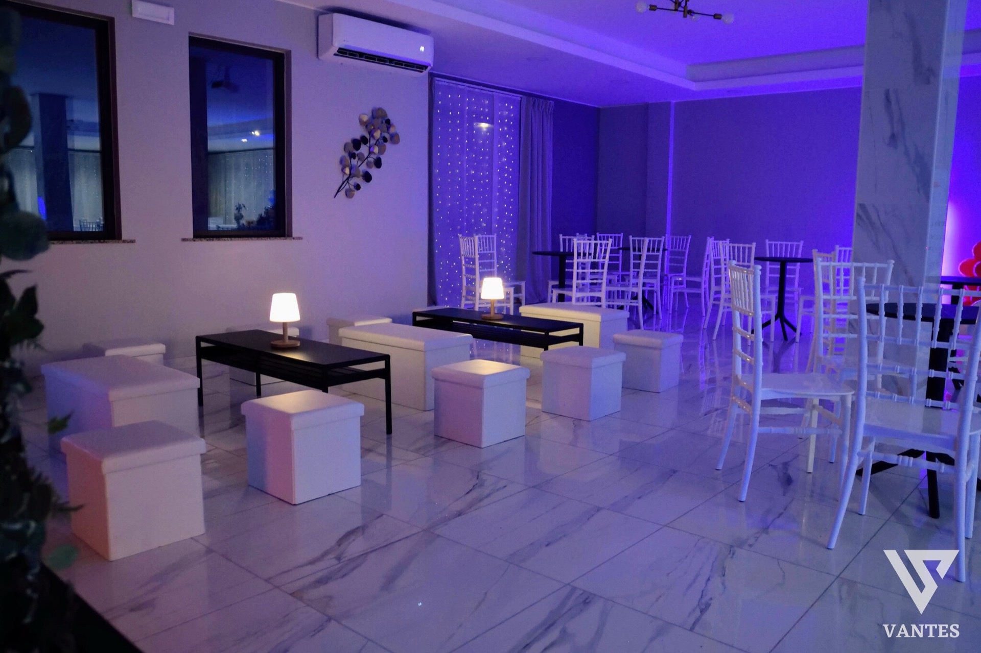 Lounge interna illuminata da luci viola con pouf bianchi, tavolini neri, sedute alte e tavoli rotondi sullo sfondo. Il pavimento lucido effetto marmo e le tende con micro-luci creano un angolo relax elegante e raccolto per il ricevimento di laurea.