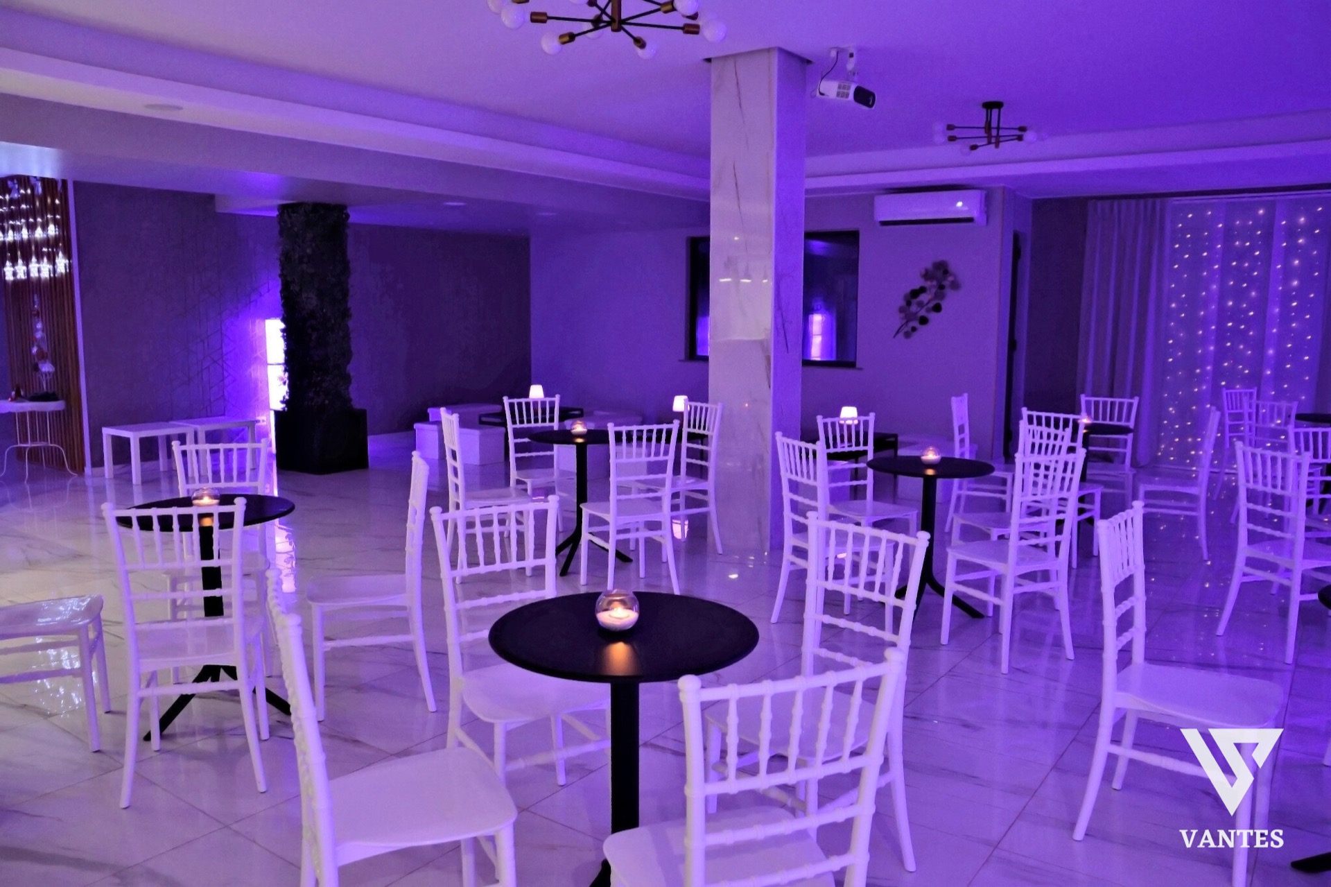 Ampia lounge interna con luce viola diffusa, tavolini rotondi neri, sedie bianche, pouf quadrati e piccole luci decorative sul fondo. L'ambiente è ordinato e minimale, pensato come area relax del ricevimento di laurea, con pavimento lucido e pareti chiare.