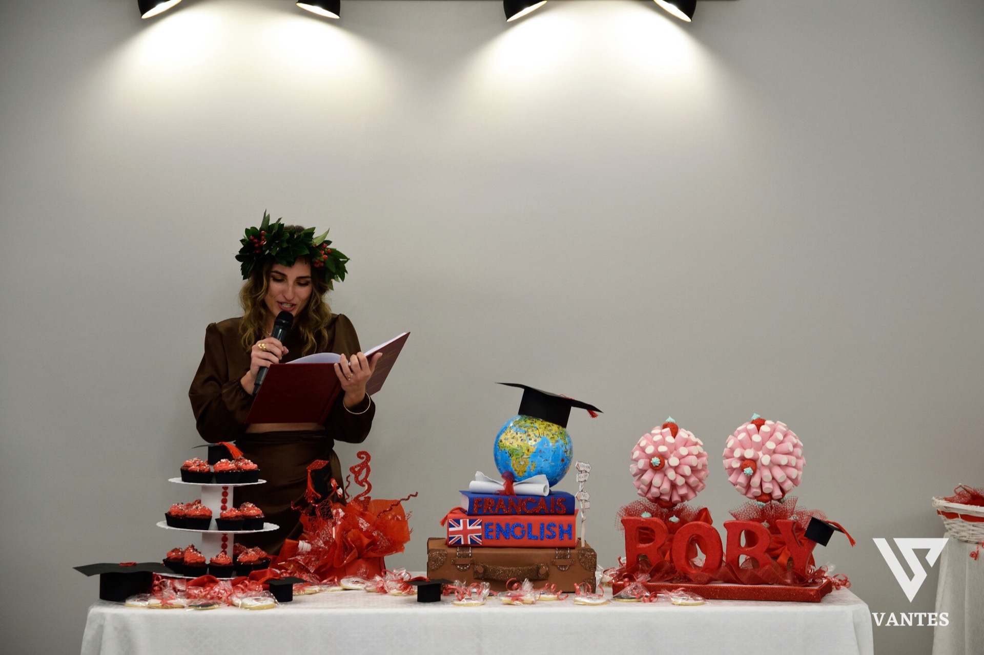 Tavolo torta per laurea con una giovane laureata che legge o parla al microfono davanti a un allestimento rosso e arancione. Sul tavolo ci sono dolci decorati, la scritta 'FRANCAIS ENGLISH' su un libro decorativo, un globo con tocco da laurea e grandi elementi con il nome della festeggiata.