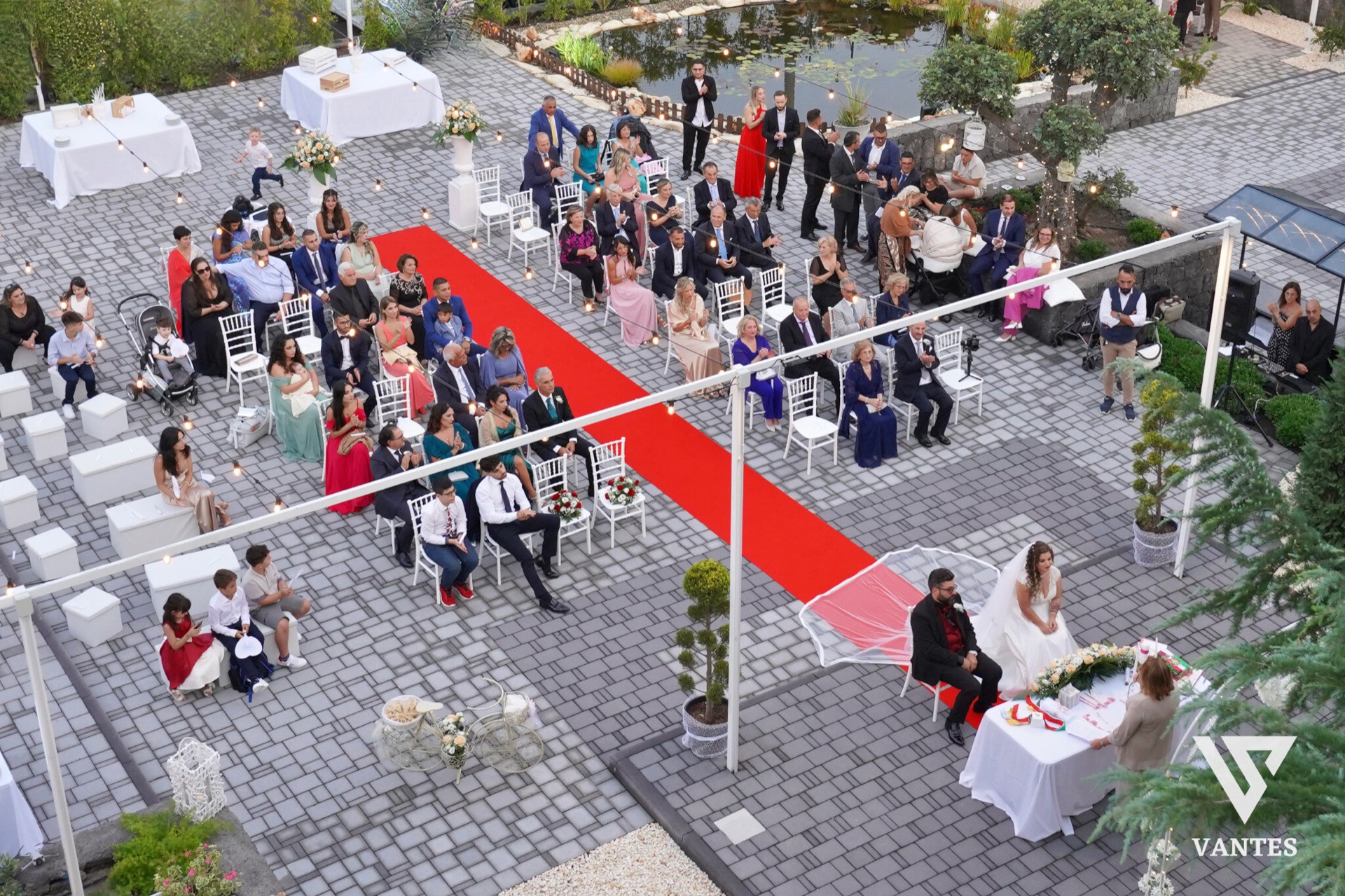 Vista dall'alto della cerimonia di matrimonio all'aperto con tappeto rosso centrale, sedie bianche disposte in più blocchi e ospiti seduti su entrambi i lati. In basso a destra si vede il tavolo degli sposi, mentre sullo sfondo compaiono il laghetto, le decorazioni floreali e i punti luce sospesi tra gli alberi.