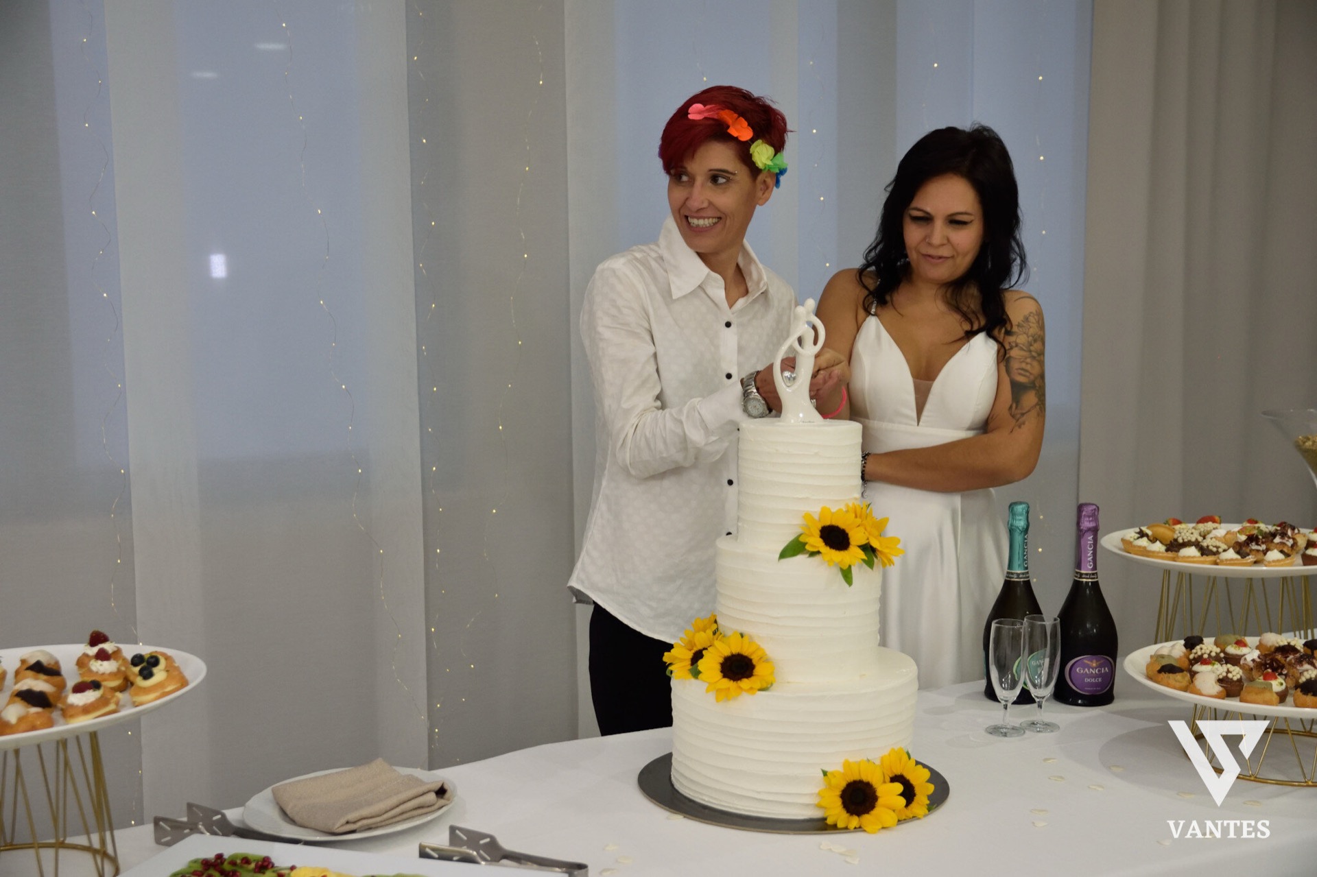 Taglio torta in sala con una torta bianca a tre piani decorata con girasoli e le due protagoniste che tengono insieme il coltello davanti al dolce. Ai lati del tavolo ci sono buffet di pasticcini, bottiglie di spumante e calici, mentre lo sfondo è composto da tende chiare e luci decorative verticali.