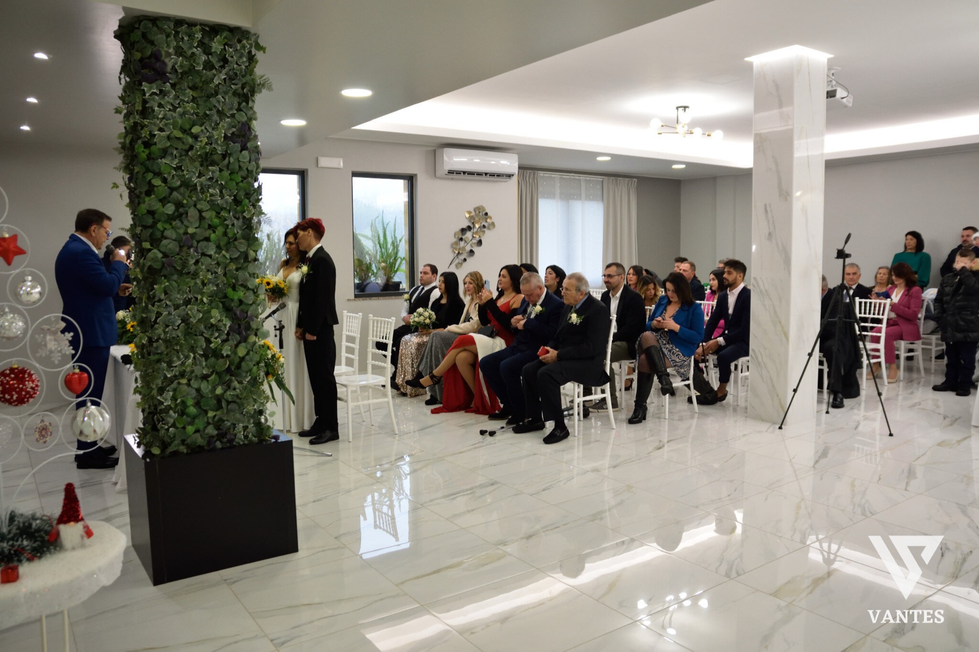 Cerimonia di matrimonio in sala con il celebrante che legge davanti agli sposi, mentre gli invitati sono seduti in file ordinate su sedie bianche. La scena è in un ambiente luminoso con pavimento lucido effetto marmo, una parete verde verticale a sinistra e un piccolo angolo decorativo sullo sfondo.