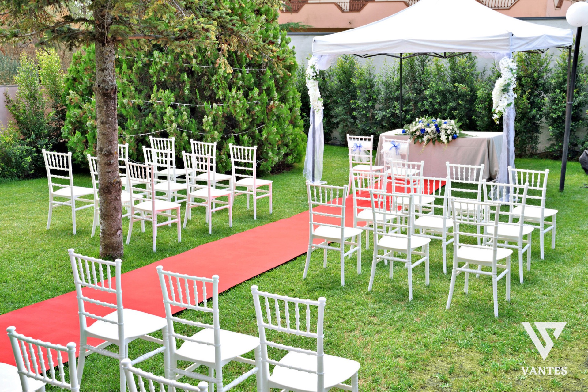 Rito di matrimonio all'aperto nel prato con tappeto rosso che conduce all'altare, sedie bianche disposte ai lati e un tavolo decorato sotto il gazebo. Sullo sfondo si vedono grandi alberi, catenarie luminose e il giardino che fa da cornice alla celebrazione.