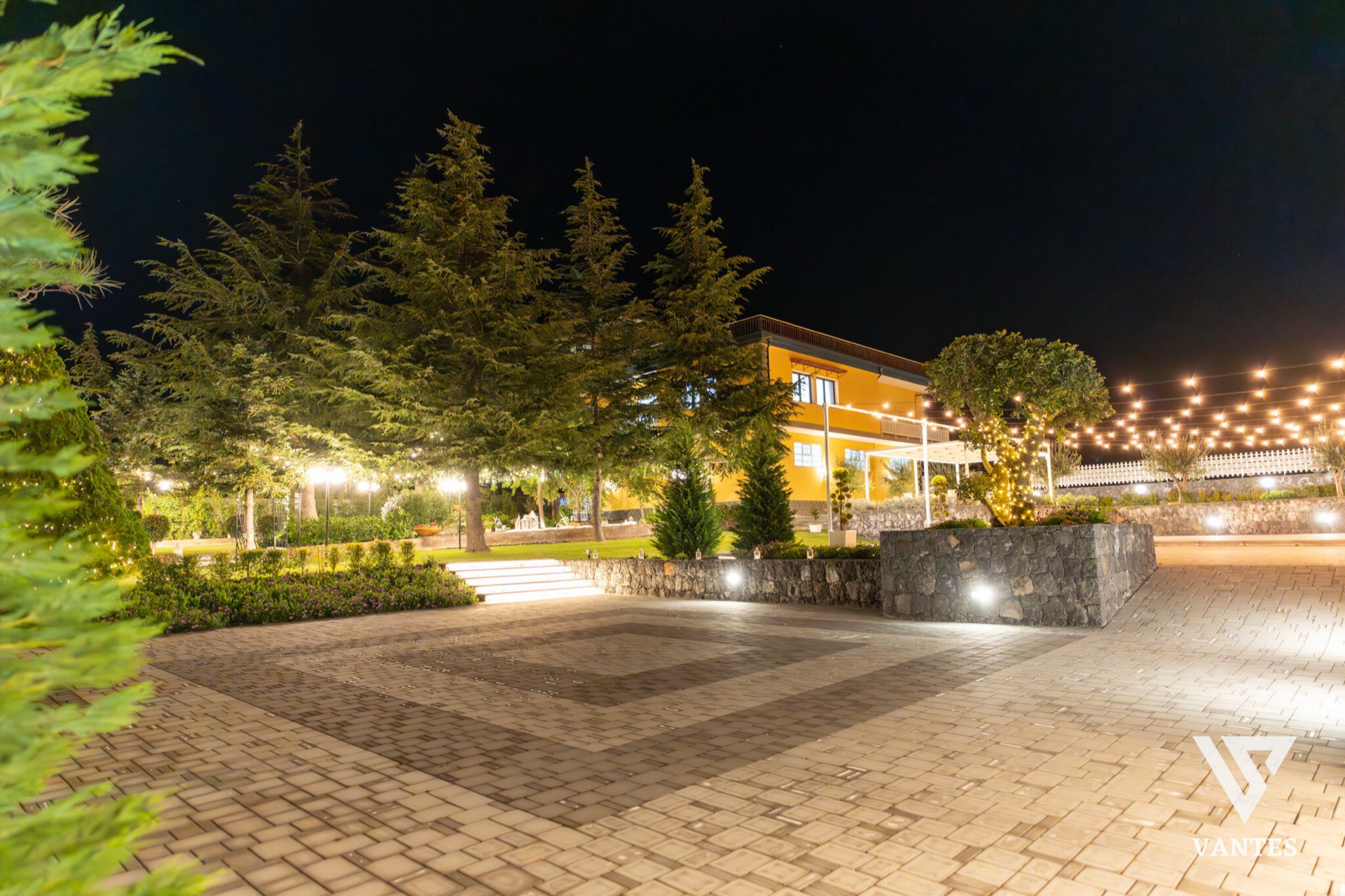 Ampia vista notturna del giardino con pavimentazione in mattoni, alberi alti illuminati dal basso, villa gialla sullo sfondo e catenarie di luci calde sospese sopra l'area esterna. La scena mostra lo spazio evento vuoto e scenografico, pronto per il ricevimento di matrimonio.