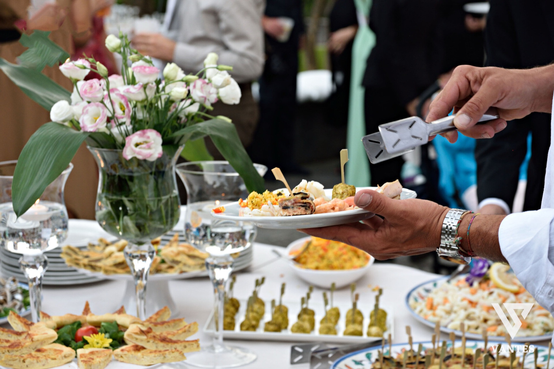 Servizio buffet con un cameriere che impiatta finger food e antipasti su un piatto bianco, usando una pinza mentre gli ospiti attendono sullo sfondo. Sul tavolo ci sono vassoi di tartine, spiedini, insalate e una composizione floreale centrale, in un momento di catering molto ravvicinato.
