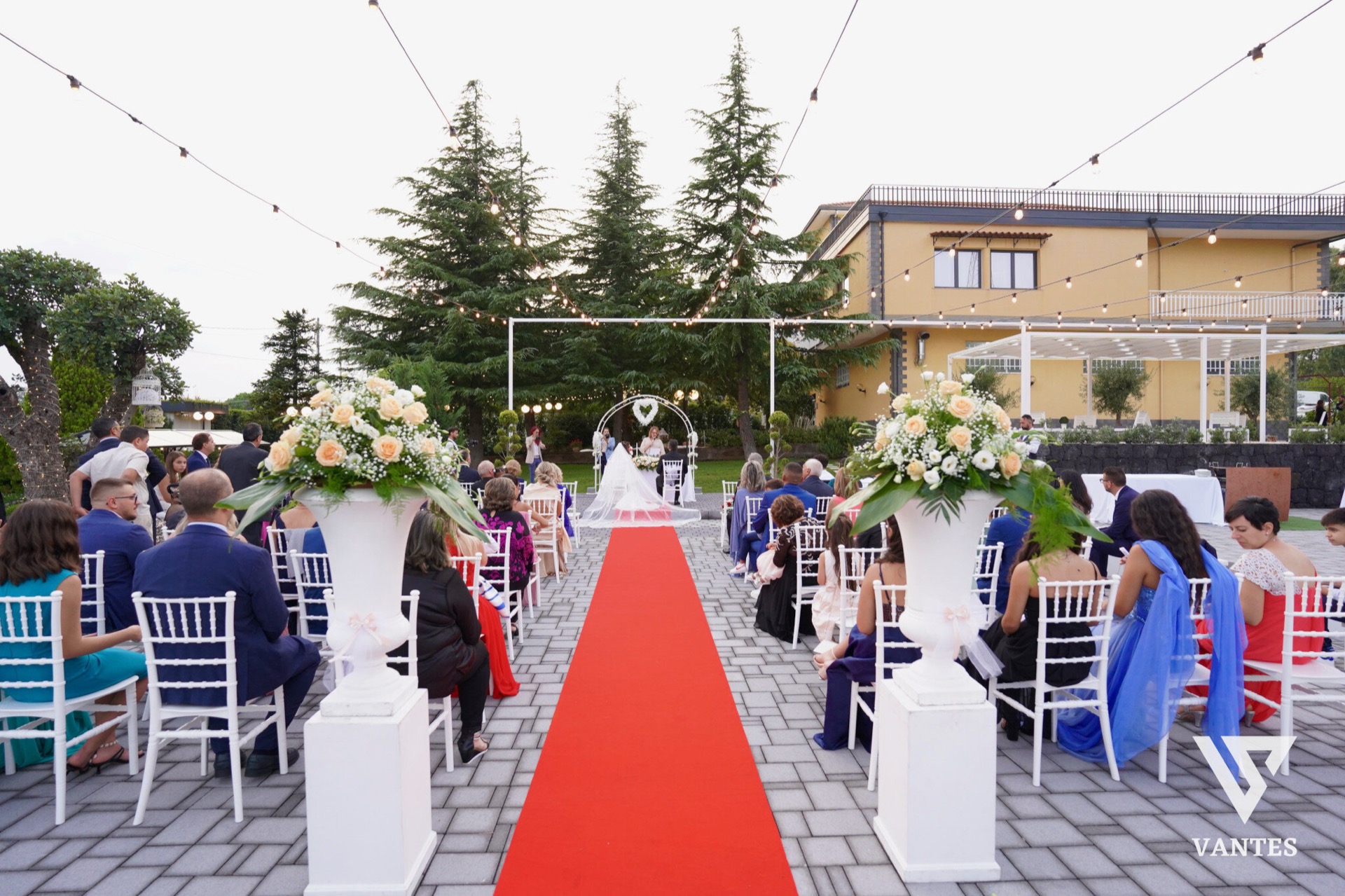 Cerimonia di matrimonio all'aperto vista frontalmente con tappeto rosso centrale, sedie bianche su entrambi i lati e due grandi vasi di fiori chiari all'ingresso del percorso. Sullo sfondo si vede l'altare con gli sposi, il giardino illuminato e la villa gialla alle spalle.