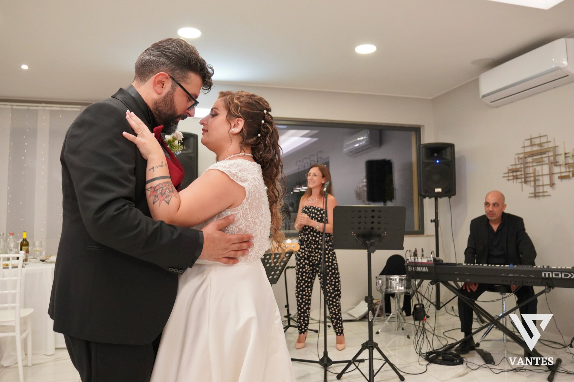 Gli sposi ballano abbracciati in sala durante il ricevimento, con lei in abito bianco e lui in completo scuro con dettaglio rosso. Sullo sfondo si vedono la cantante, il tastierista, i microfoni e le casse della band, in un momento intimo e musicale della festa.