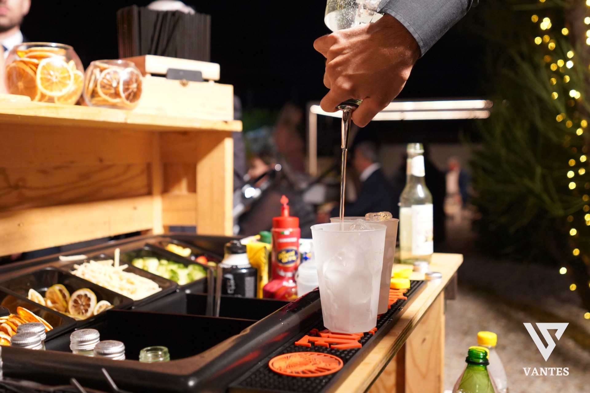 Cocktail station all'aperto di sera con un bartender che versa un drink in un bicchiere con ghiaccio, bottiglie di liquori e strumenti da bar disposti sul banco di legno. Sul lato sinistro si vedono vaschette con agrumi essiccati e altri ingredienti per la preparazione, mentre sullo sfondo brillano le luci del giardino.