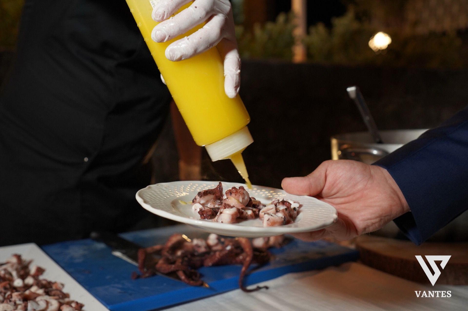 Primo piano di un piatto bianco con polpo tagliato a pezzi e una salsa gialla versata dall'alto da un flacone in mano al cuoco. Sul tavolo di servizio si vedono altre porzioni di polpo, una superficie da lavoro blu e il gesto preciso del plating durante il ricevimento di matrimonio.