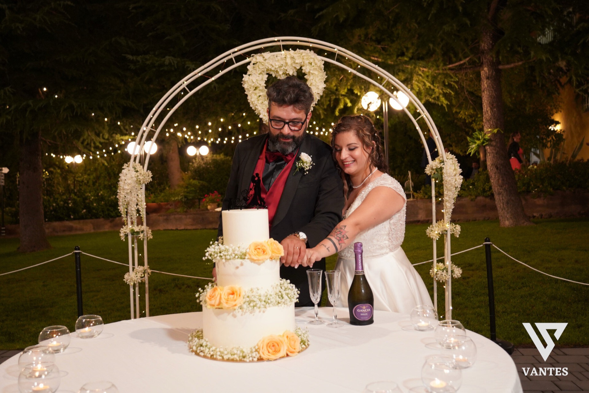 Sposi che tagliano una torta nuziale bianca a tre piani decorata con rose gialle e gypsophila, sotto un arco metallico fiorito in giardino di notte. Sul tavolo ci sono calici, una bottiglia di spumante e piccole candele, mentre dietro brillano le luci calde sospese tra gli alberi.