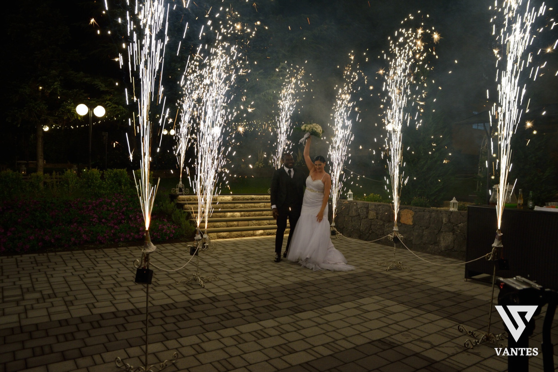 Gli sposi posano al centro del piazzale notturno circondati da alte fontane luminose che sparano scintille verso l'alto. Il marito è in abito scuro e la sposa in abito bianco solleva il bouquet, mentre sullo sfondo si vedono i gradini illuminati e le aiuole del giardino.