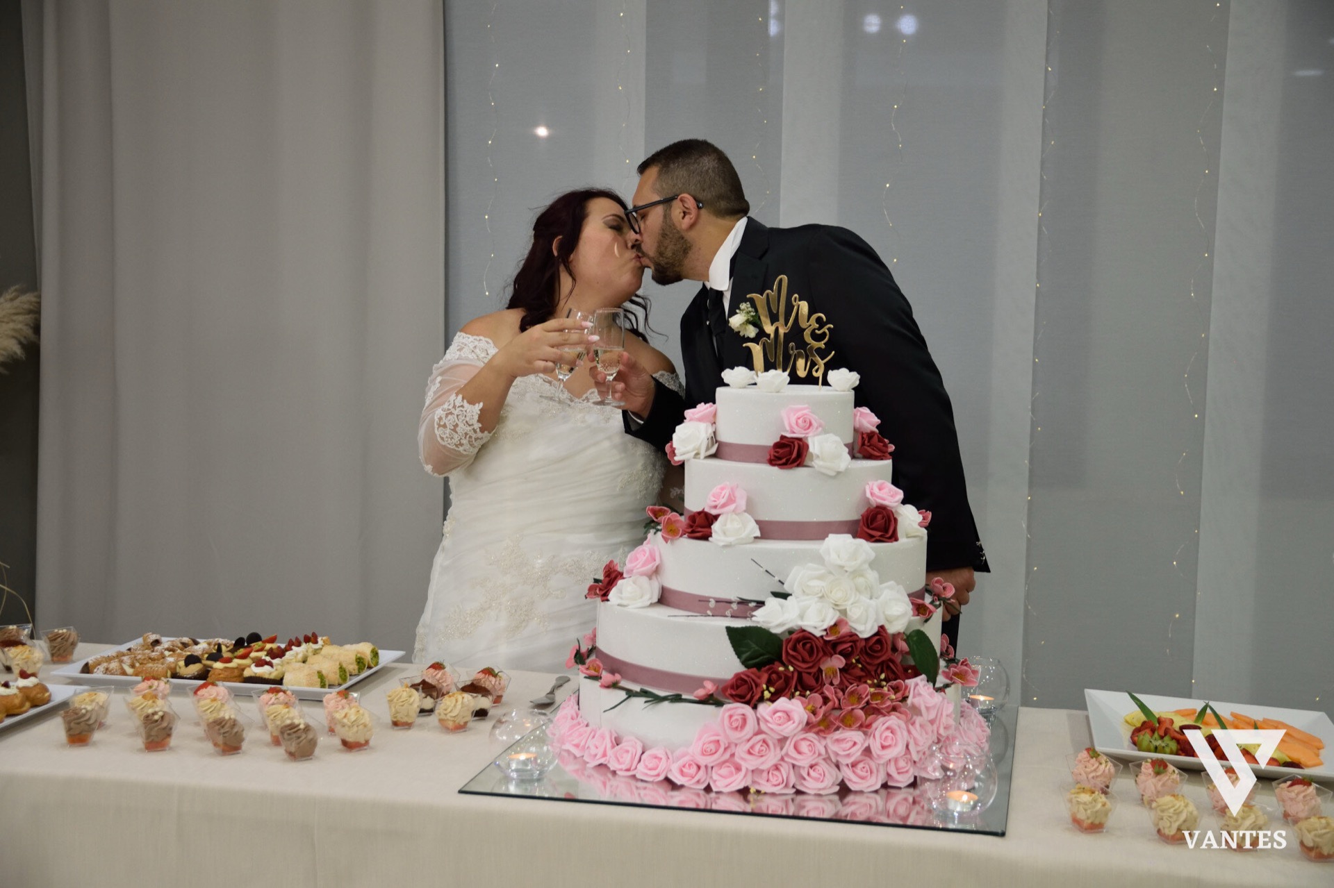 Taglio torta in sala con gli sposi che si baciano accanto a una torta alta decorata con rose rosa, rosse e bianche, su una base piena di petali rosa. Sul tavolo ci sono vassoi di dolci, bicchieri con candele e una bottiglia di spumante, mentre la parete chiara sullo sfondo è illuminata da piccoli punti luce.