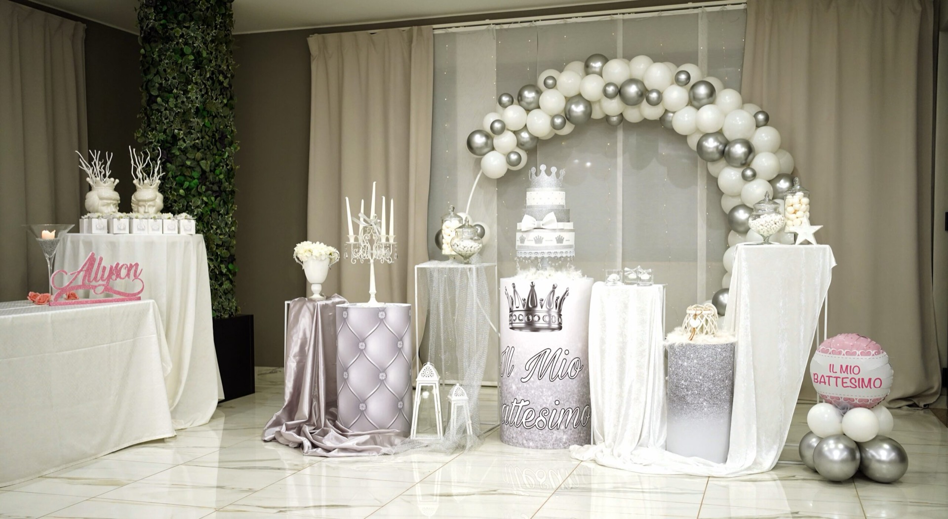 Allestimento battesimo in sala con grande arco di palloncini bianchi e argentati, torta centrale a più piani, colonne decorative rivestite in tessuto chiaro e tavoli confettata coordinati ai lati. Sullo sfondo si vedono il fondale con il nome della festeggiata, candele decorative e un ambiente elegante con tende e pavimento lucido.