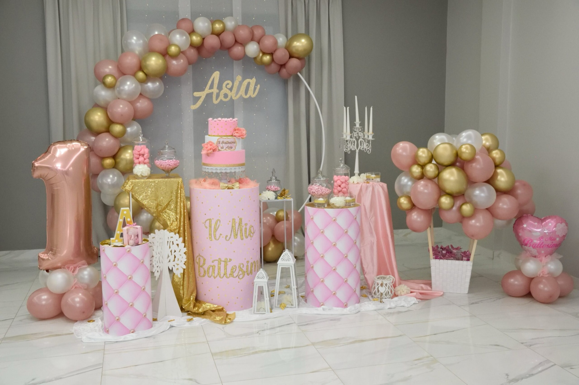 Allestimento scenografico per battesimo in sala con grande arco di palloncini rosa, oro e bianchi, numero 1 metallico, nome 'Asia' sul fondale e torta centrale a più piani in rosa e oro. Ai lati ci sono confettata, candele decorative, piccoli tavoli rivestiti in tessuto glitter oro e composizioni coordinate di palloncini.