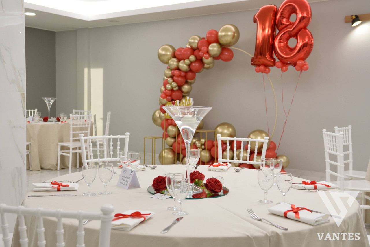 Allestimento interno per diciottesimo con grande arco di palloncini rossi e oro, numero 18 metallico rosso e tavoli rotondi apparecchiati con calici e tovaglioli legati da nastri rossi. Al centro del tavolo c'è un centrotavola con rose rosse e un ampio calice martini, in un ambiente elegante e luminoso.