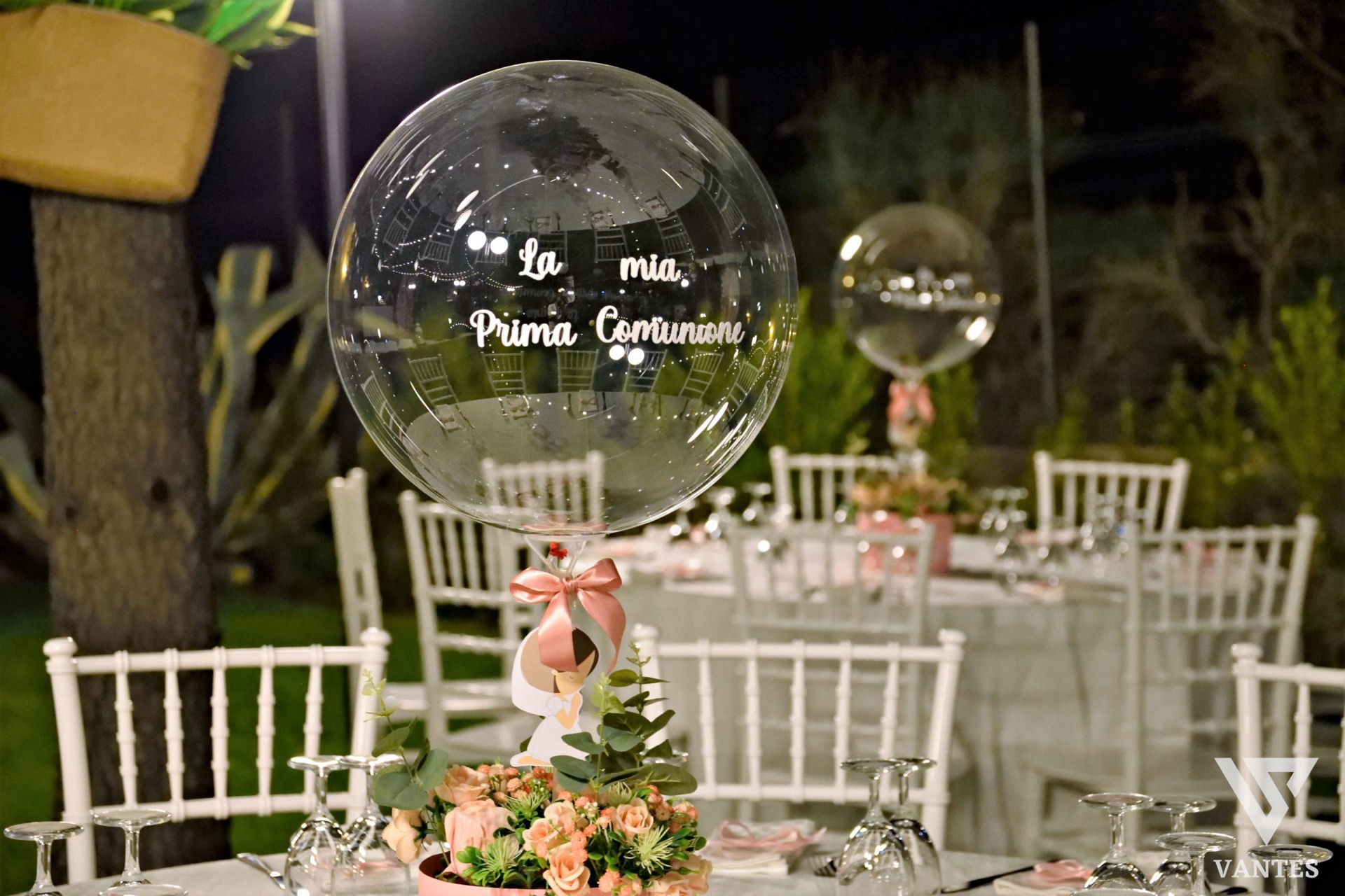 Tavolo allestito all'aperto per la Prima Comunione con palloncino trasparente personalizzato 'La mia Prima Comunione' in primo piano, nastro rosa e composizione floreale delicata alla base. Sullo sfondo si vedono tavoli rotondi apparecchiati con sedie bianche, calici rovesciati e l'atmosfera serale del giardino.