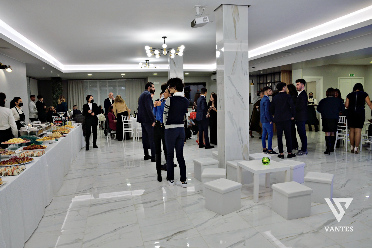 Sala interna per apericena di laurea con lungo buffet laterale, tavoli con antipasti e ospiti in piedi al centro dell'ambiente luminoso. I pilastri chiari, il pavimento lucido e l'illuminazione uniforme danno alla scena un aspetto moderno e ordinato.