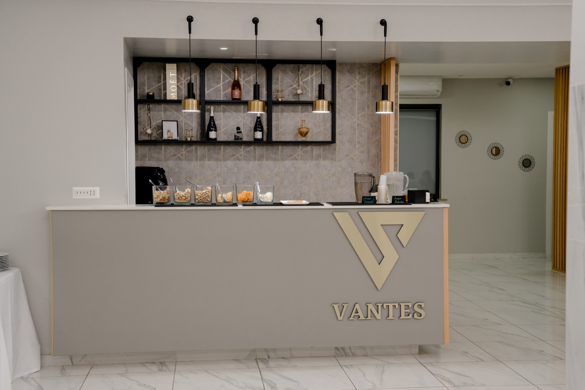 Banco bar interno della sala con linee minimal, bottiglie esposte su scaffalature retroilluminate e piccoli snack e bevande sul piano. L'ambiente è luminoso, ordinato e pensato per il servizio drink durante l'evento.