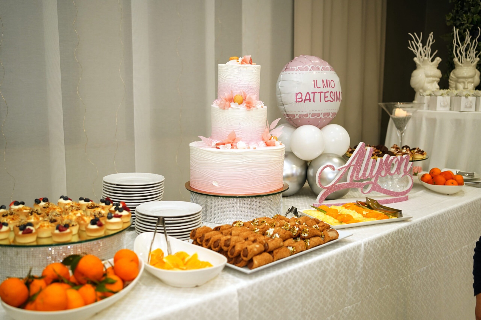 Buffet dolci per battesimo con torta rosa a più piani al centro, pallone decorativo con la scritta 'Il mio battesimo', piatti di pasticcini, frutta fresca e torte monoporzione disposti su una tavola elegante. Sul fondo si vedono anche i dettagli decorativi coordinati in bianco e rosa.