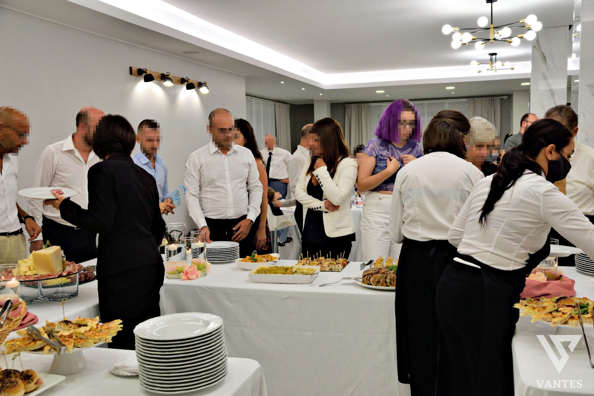 Sala buffet molto affollata con numerosi ospiti in piedi attorno ai tavoli di servizio e personale in divisa bianca e nera che distribuisce piatti. I tavoli sono coperti di antipasti, vassoi caldi, finger food, piatti impilati e piccole decorazioni floreali, in un ambiente moderno e luminoso.