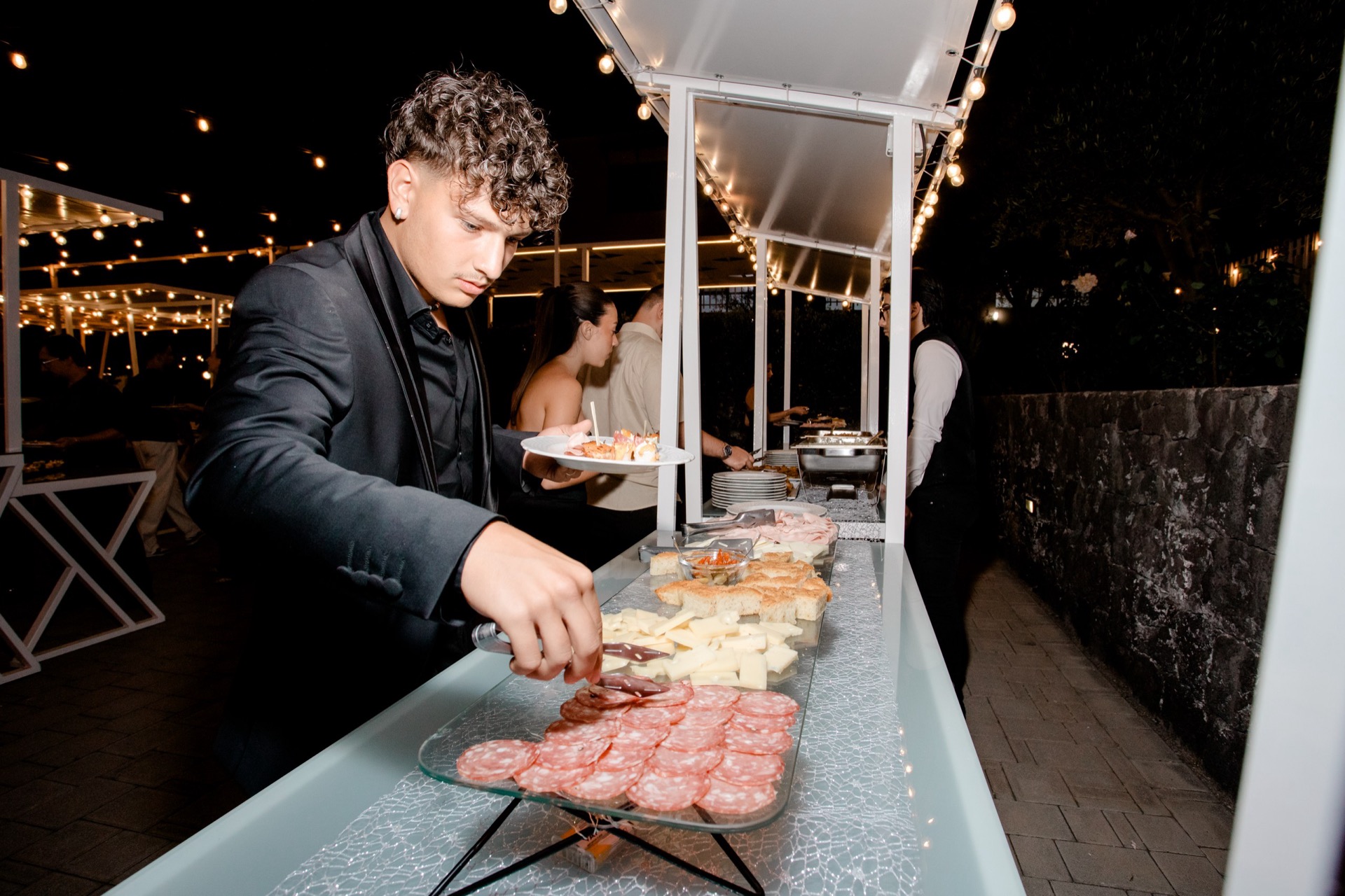 Buffet esterno serale con salumi, formaggi, panini e piccoli finger food disposti in fila sotto le luci calde della pergola. Un cameriere in abito scuro serve i piatti mentre gli ospiti si muovono lungo il banco all'aperto.