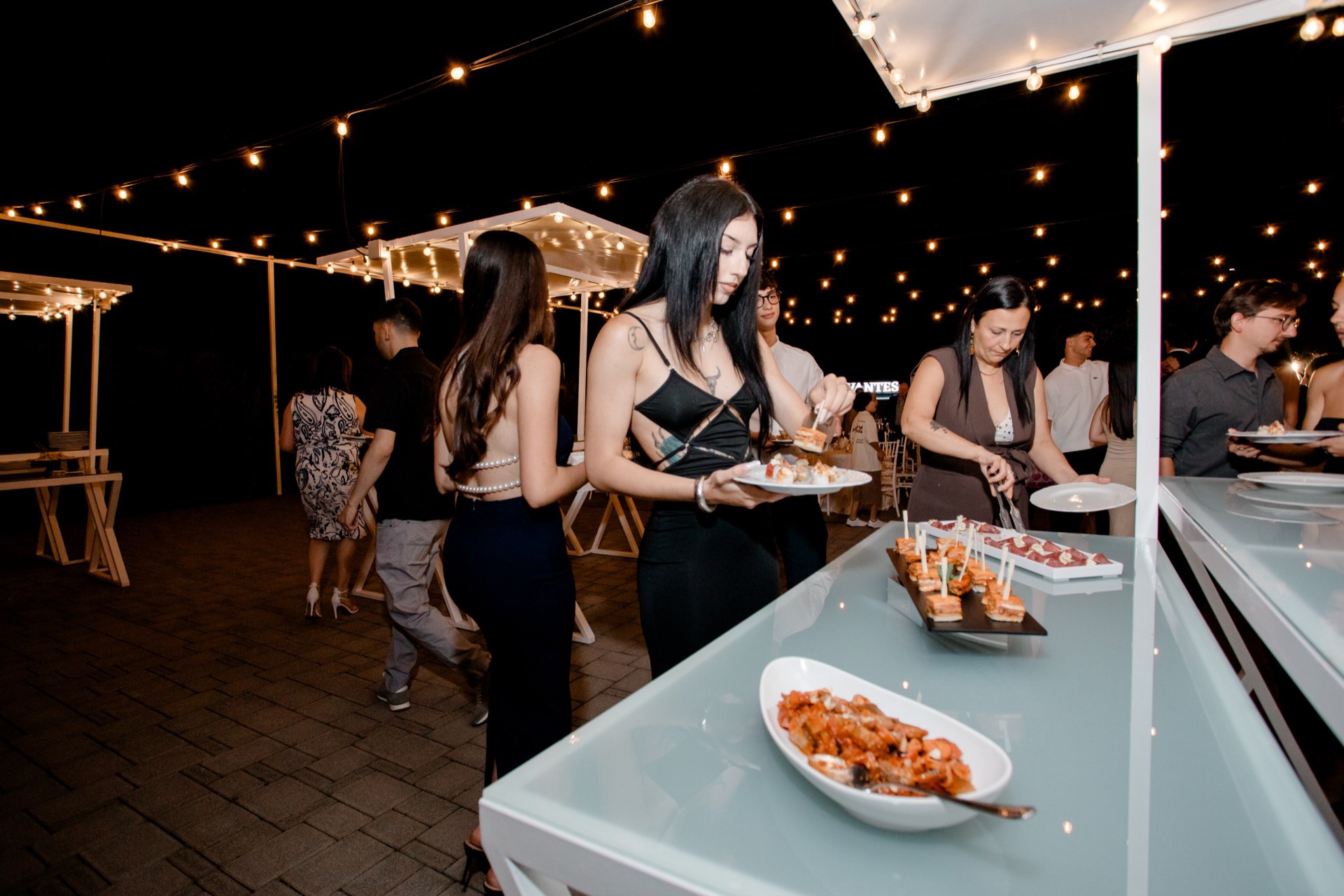 Buffet notturno all'aperto con un cameriere che serve salumi e formaggi da un lungo banco illuminato da catenarie calde. Sul tavolo ci sono vassoi di affettati, cubi di formaggio, pane, antipasti e piatti pronti, mentre gli ospiti si avvicinano lungo il percorso esterno.