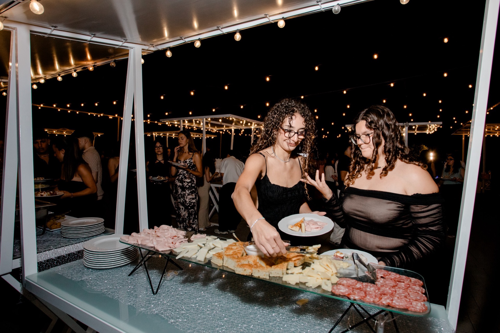 Buffet esterno notturno con due ospiti che si servono davanti al banco di salumi, formaggi, pane e antipasti, sotto le luci calde sospese nel giardino. Sullo sfondo si intravedono altri invitati e il servizio allestito sotto le pergole illuminate.