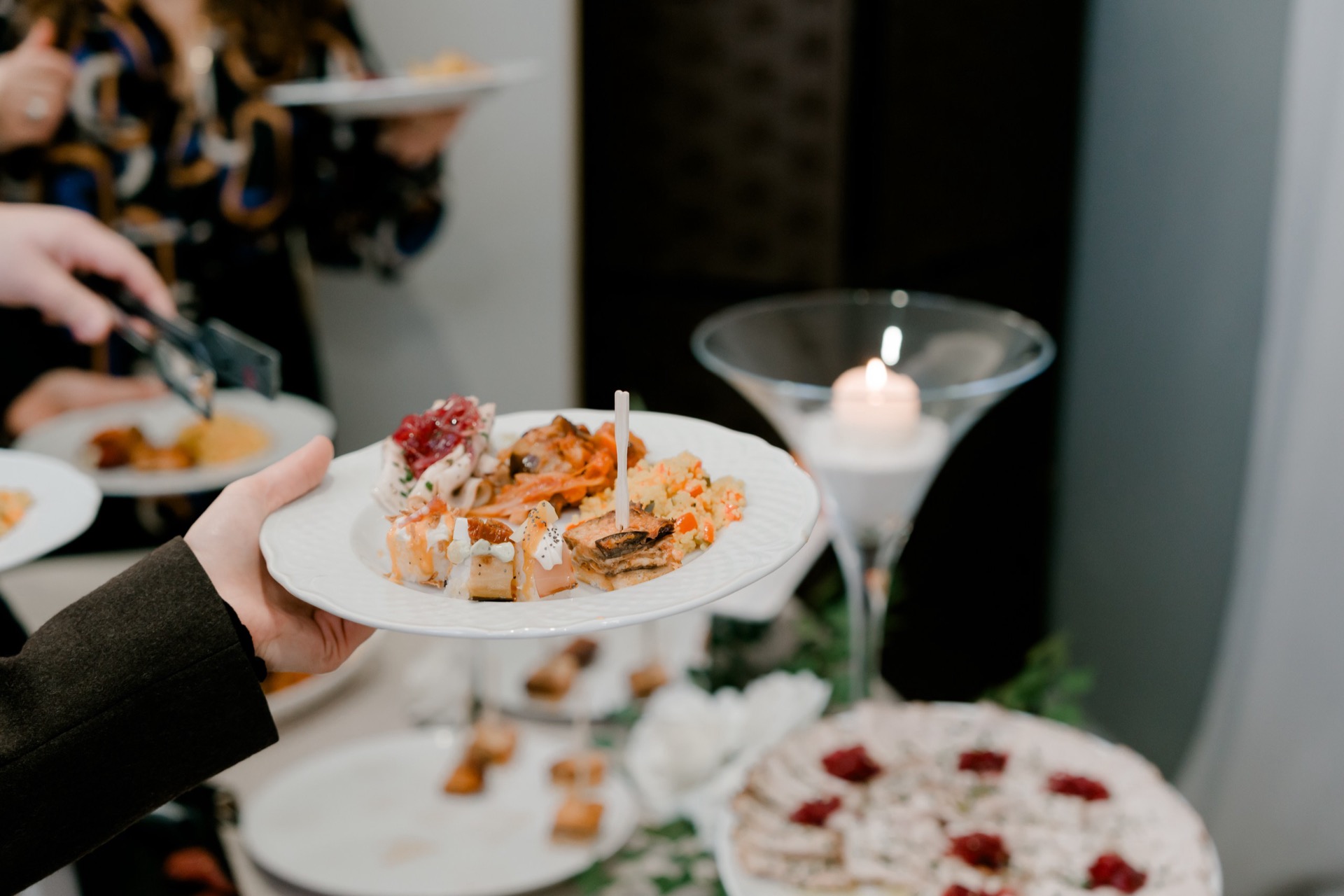 Primo piano di un piatto con assaggi di buffet servito in sala, composto da piccoli bocconi salati, una porzione di riso o cereali e altri finger food vari. Sul lato si vede un grande bicchiere martini con candela, mentre sullo sfondo altri ospiti ricevono i piatti.